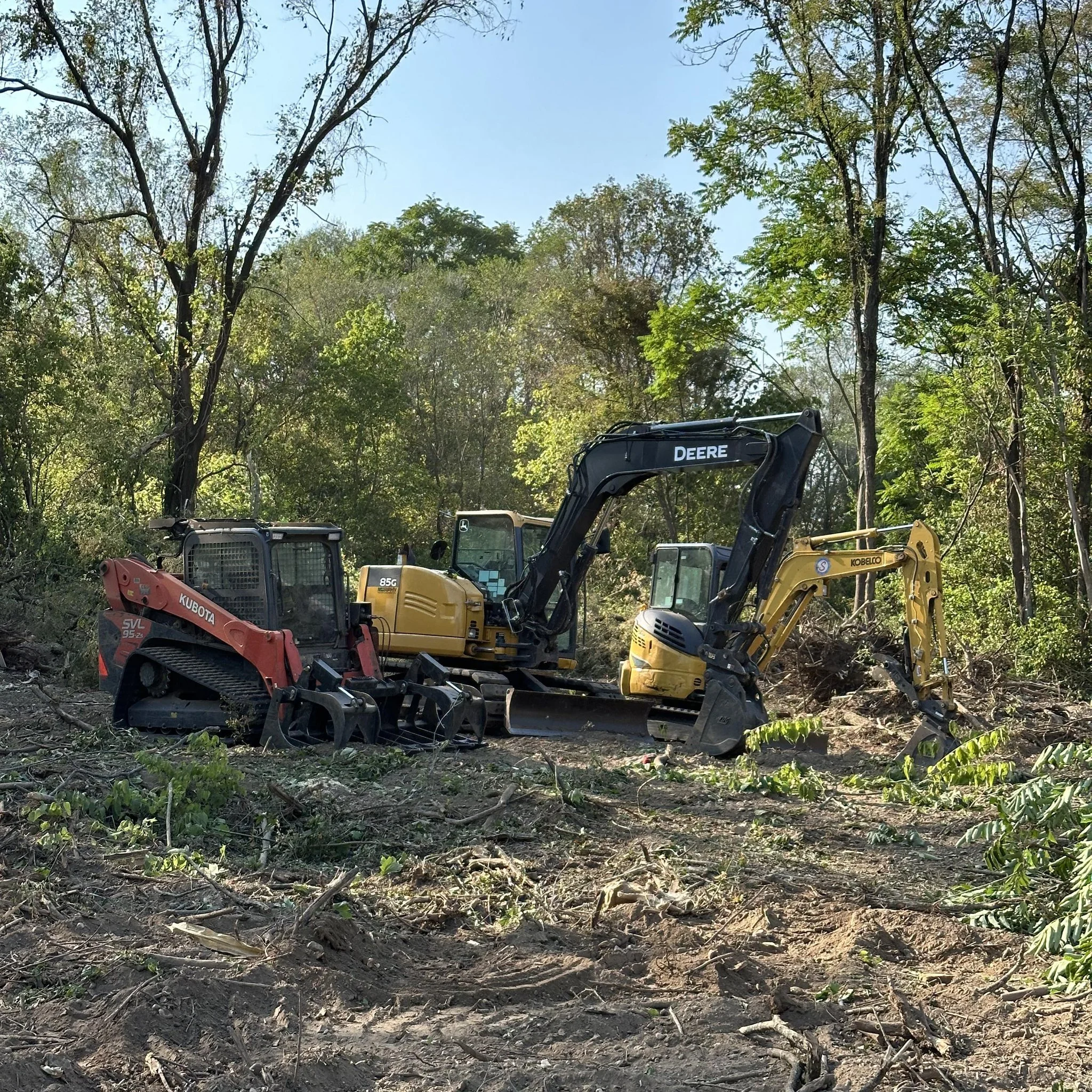 Three construction vehicles, including a red Kubota compact track loader, a yellow Deere excavator, and a yellow Kobelco mini excavator, working together to clear land in a wooded area with trees, dirt, and debris. C.C Land Solutions