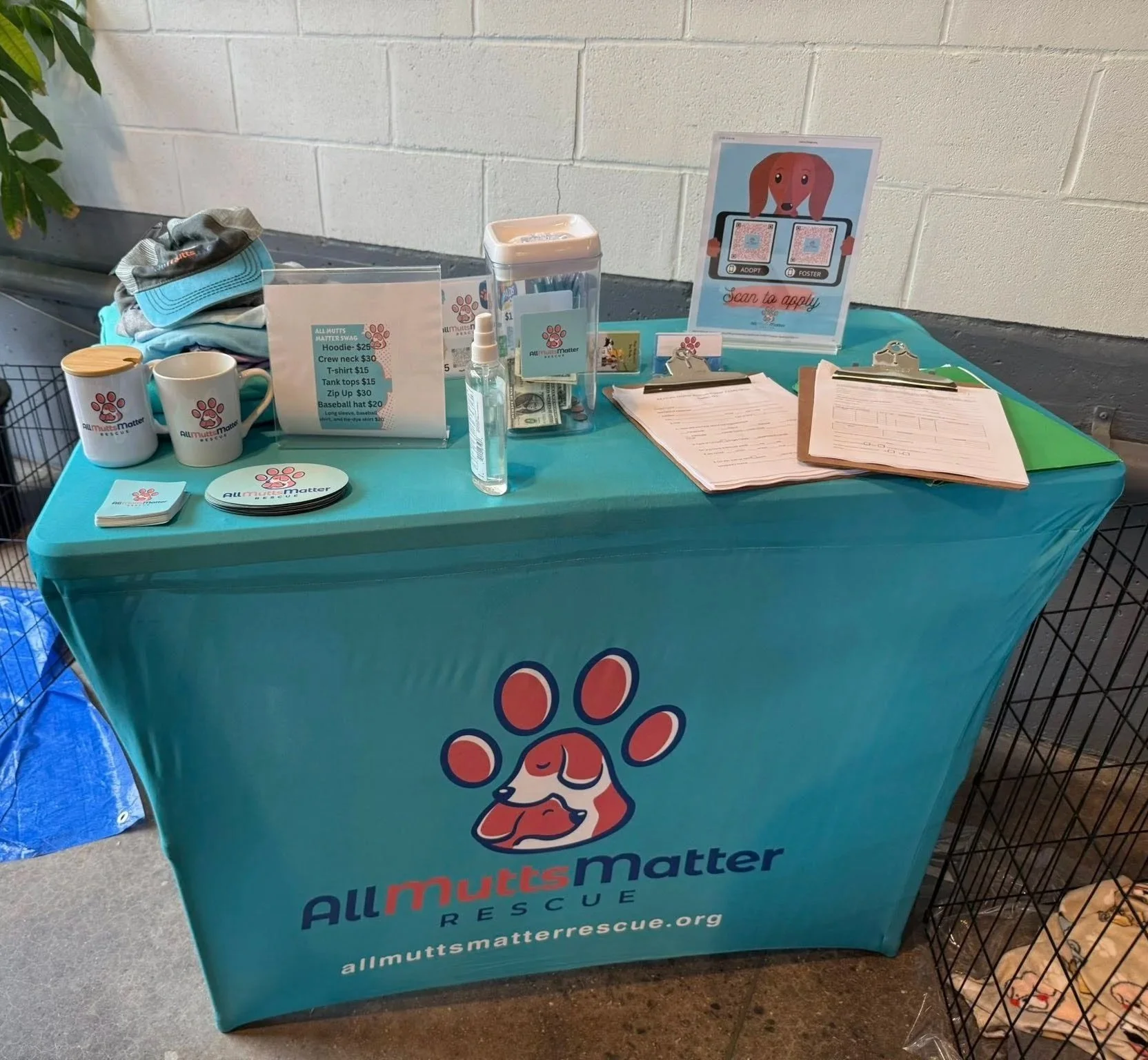 A table draped with a teal cloth displaying All Mutts Matter Rescue promotional items, including mugs, buttons, and informational brochures, with a sign promoting dog adoption and fostering.