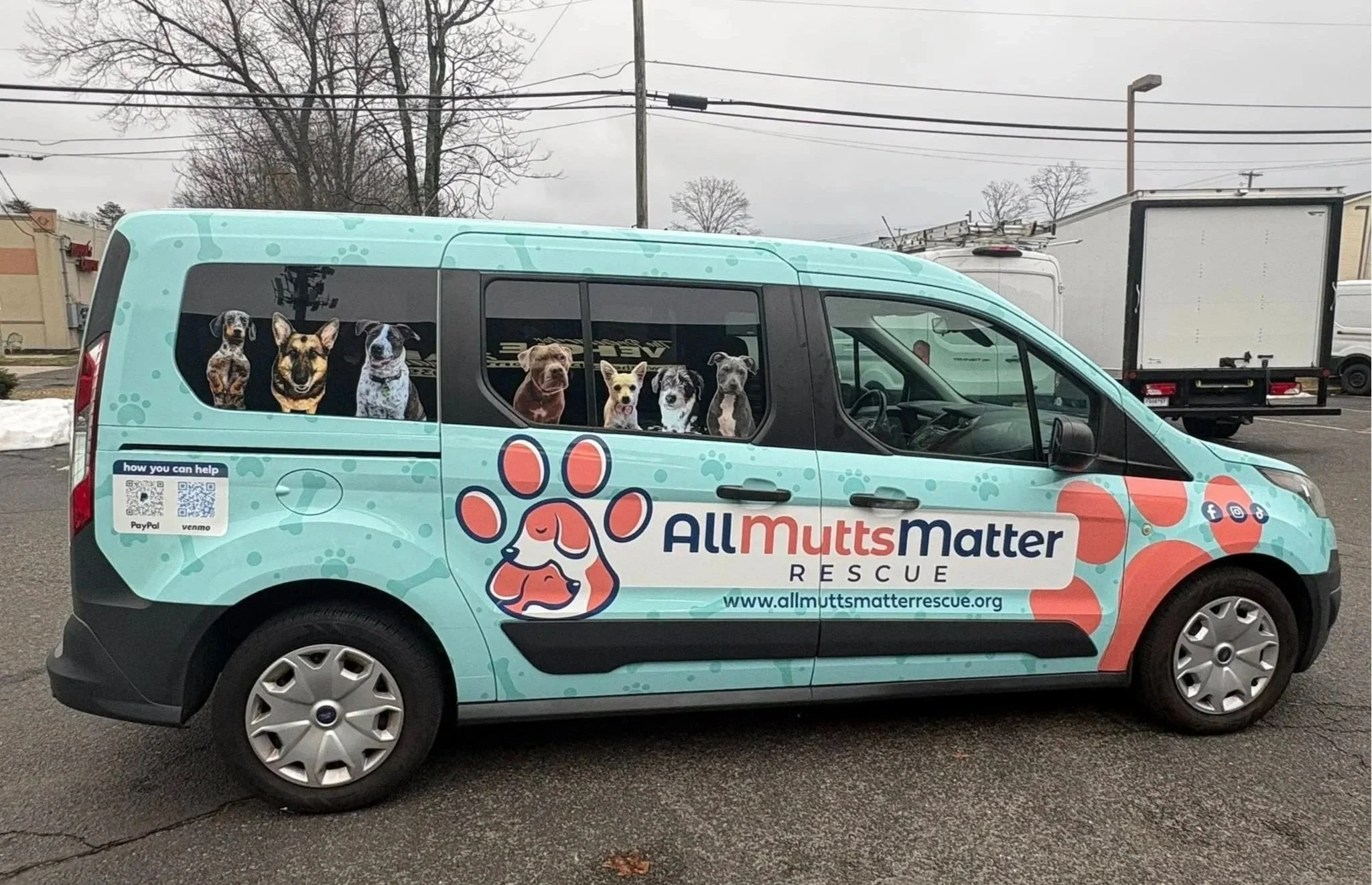 A rescue vehicle with All Mutts Matter Rescue logo and photos of several dogs on the side, parked on an overcast day in a parking lot.