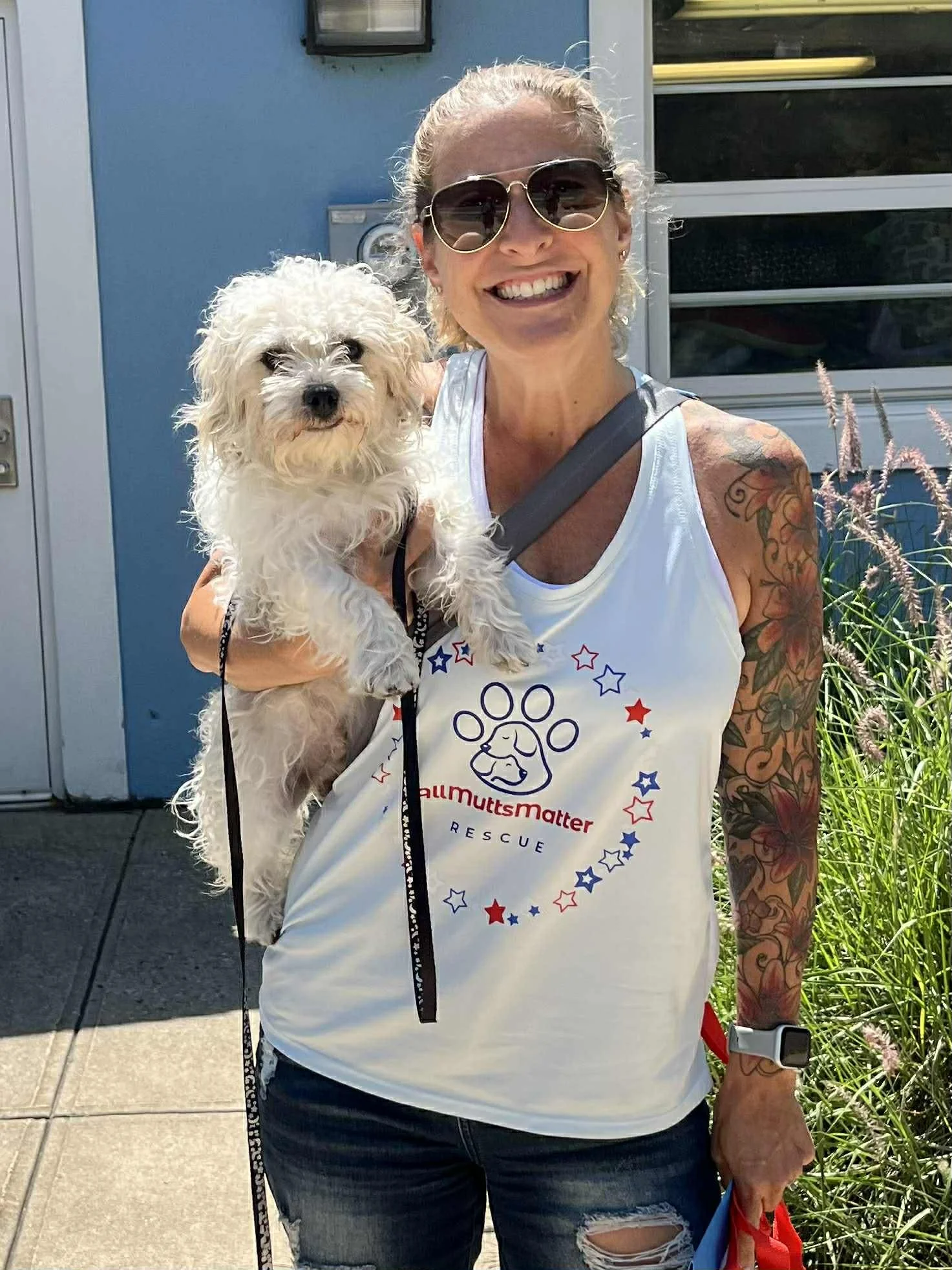 A smiling woman with sunglasses, holding a small fluffy dog, standing outdoors near a blue building with plants.