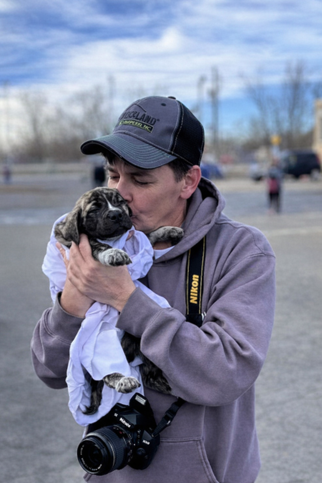 A man wearing a gray hoodie and a black cap kisses a small puppy that he is holding in his arms. The puppy is dressed in a white shirt, and the man has a Nikon camera hanging around his neck. They are outdoors, with a parking lot and some blurred people and trees in the background, under a partly cloudy sky.
