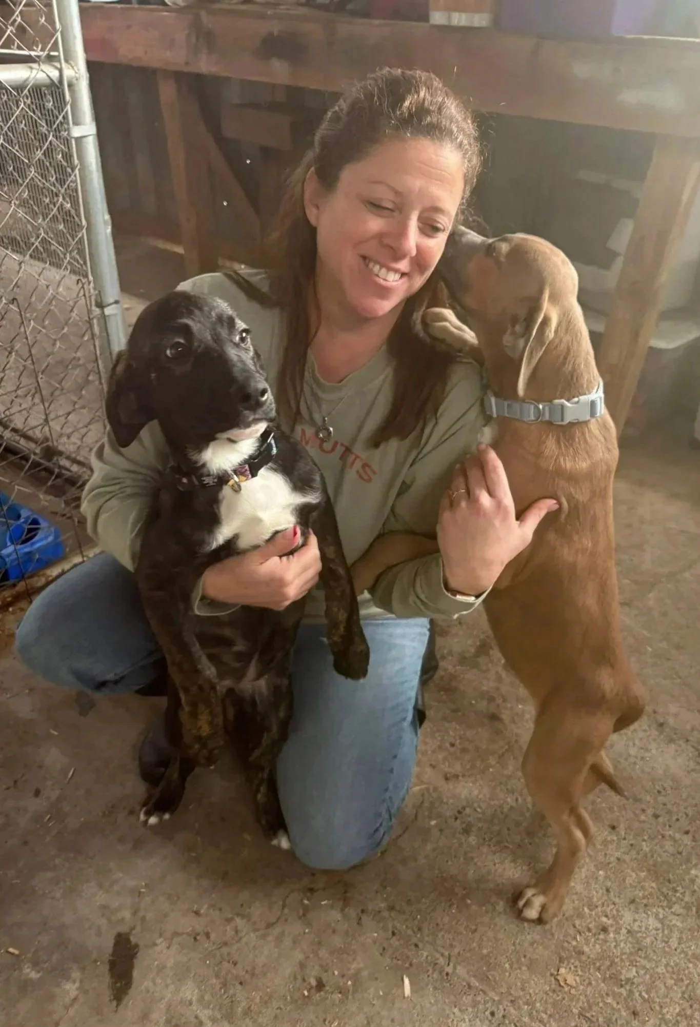 A woman kneeling on the ground smiling while holding two dogs, one black and white and the other brown, in a rustic indoor setting.
