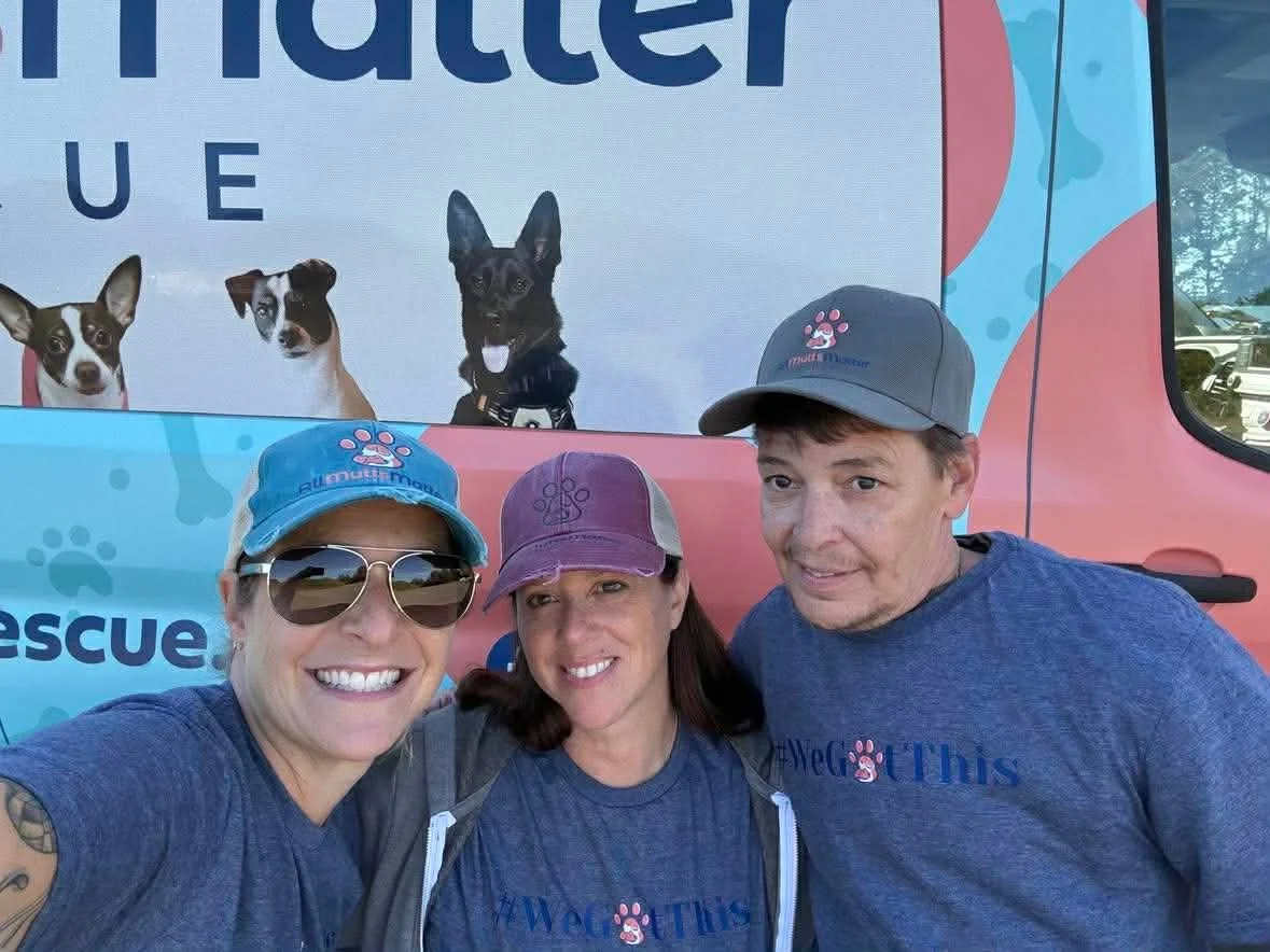 Three people taking a selfie in front of rescue dog marketing in front of a rescue vehicle, wearing matching blue shirts and caps, smiling at the camera.