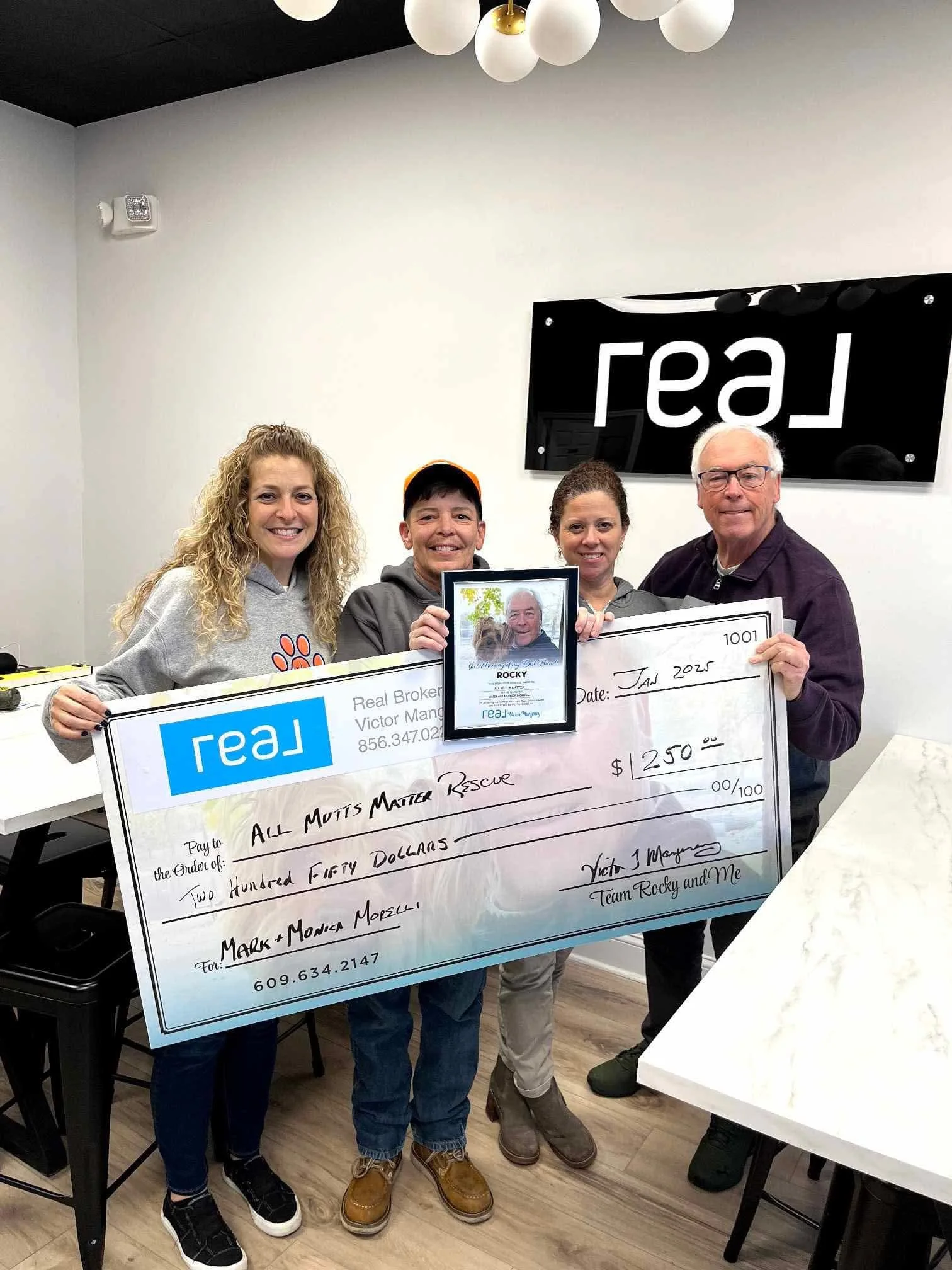 Group of four people holding an oversized check and a framed photo in an office. The check is made out for $250 to All Mutts Matter Rescue. The photo features a man outdoors. The background has a black wall with a white sign that says 'GSR'.