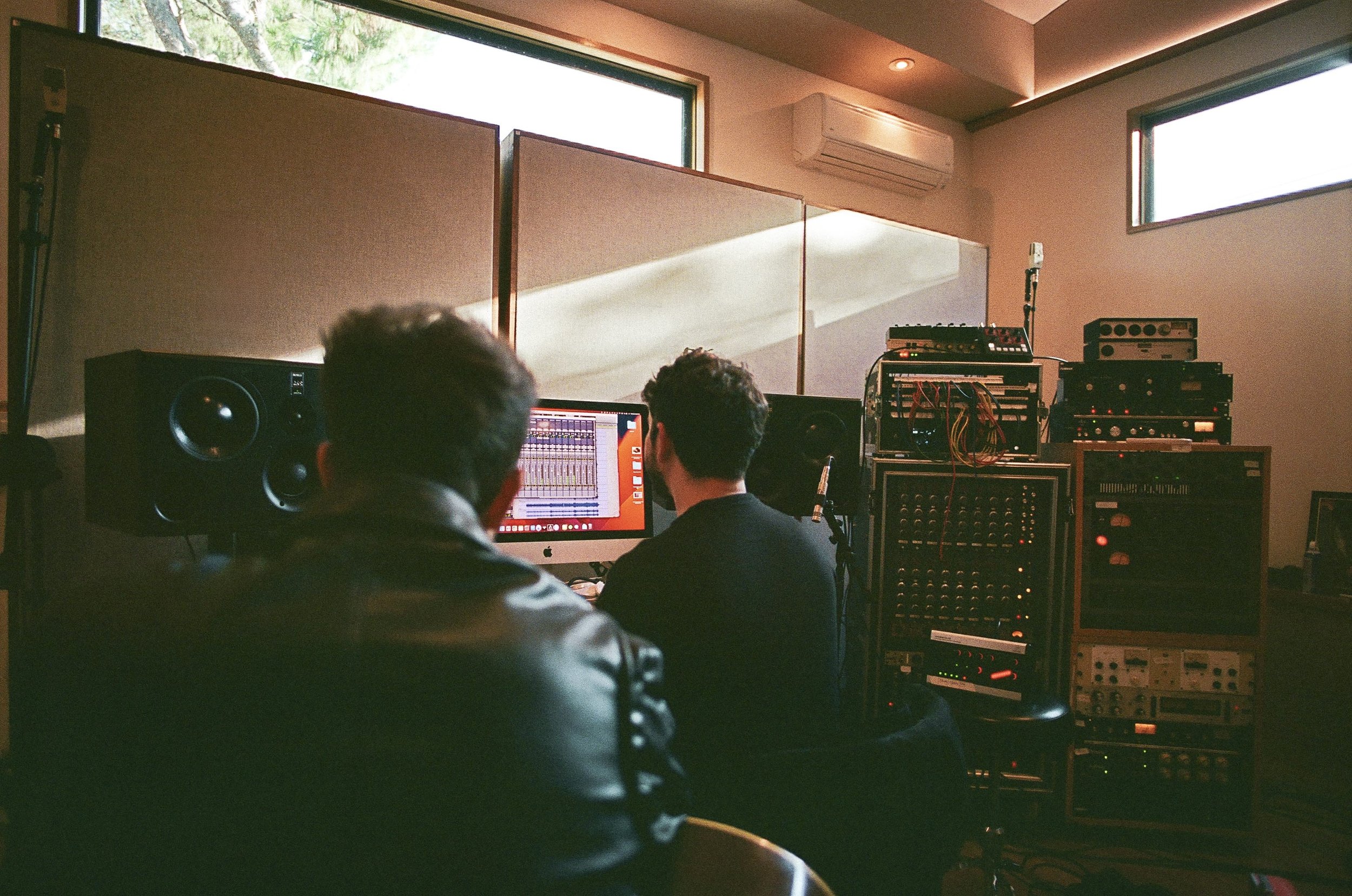 A recording studio with two people working at a computer and audio equipment. Large speakers, mixing console, and soundproofing panels are visible. Natural light coming through windows.