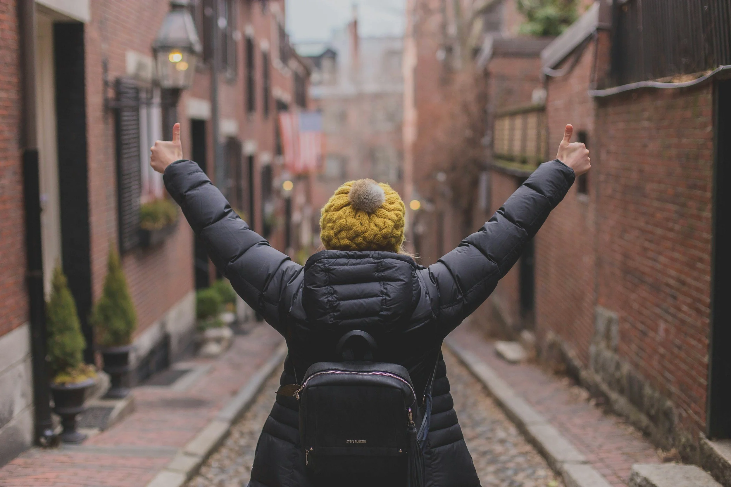 Person in a dark puffer jacket and yellow knit beanie with pom-pom, facing away, with arms raised giving a thumbs-up gesture through an alley with brick buildings.