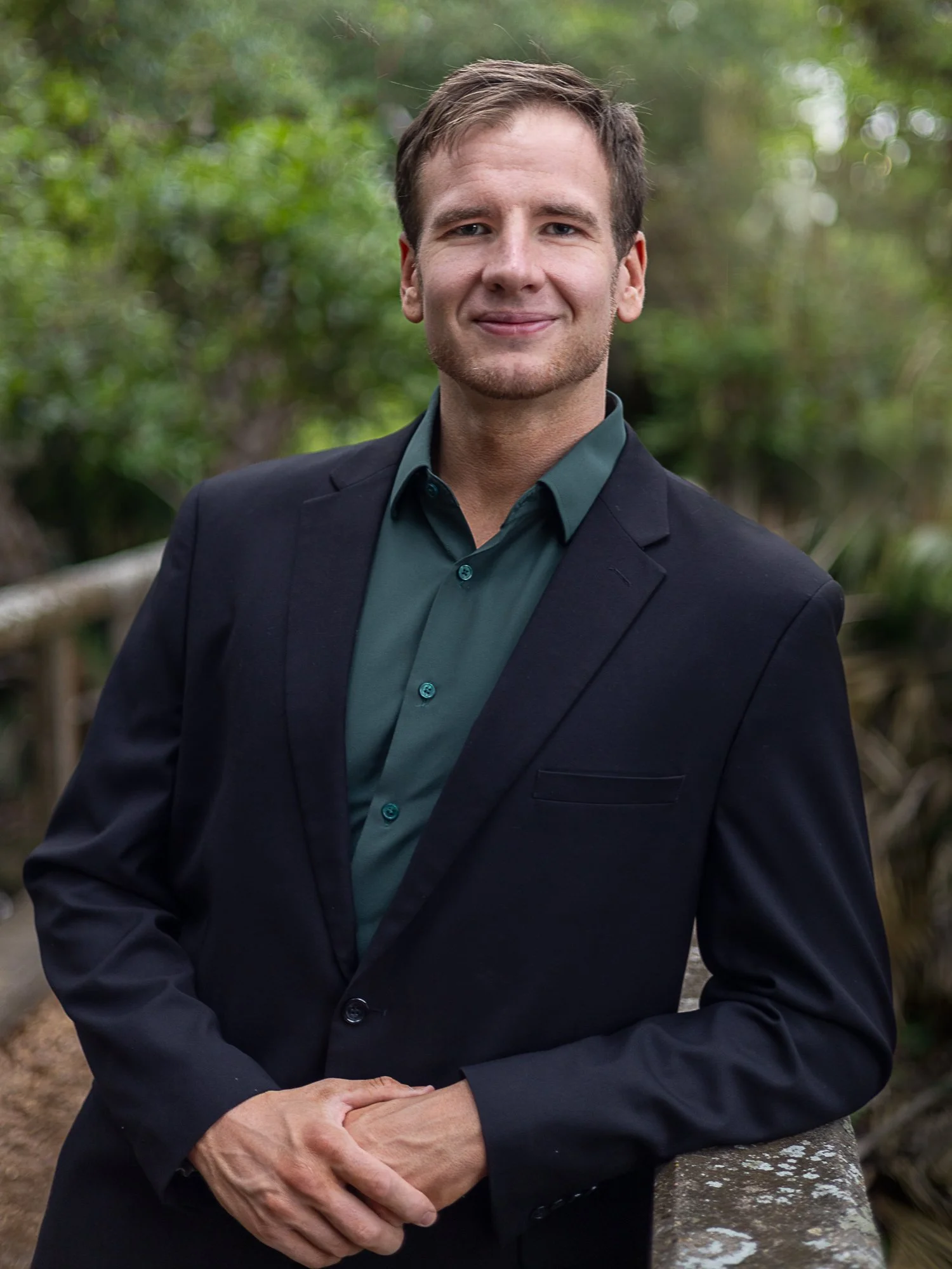 A young man wearing a dark blazer and green shirt, standing outdoors with a blurred green background, smiling slightly.