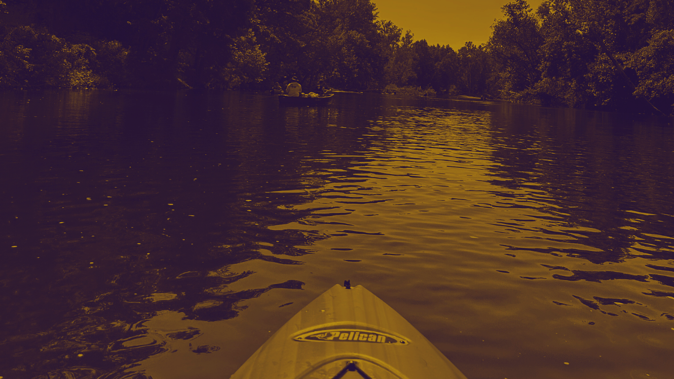 View from a kayak labeled Pelican on a river surrounded by trees on a sunny day.