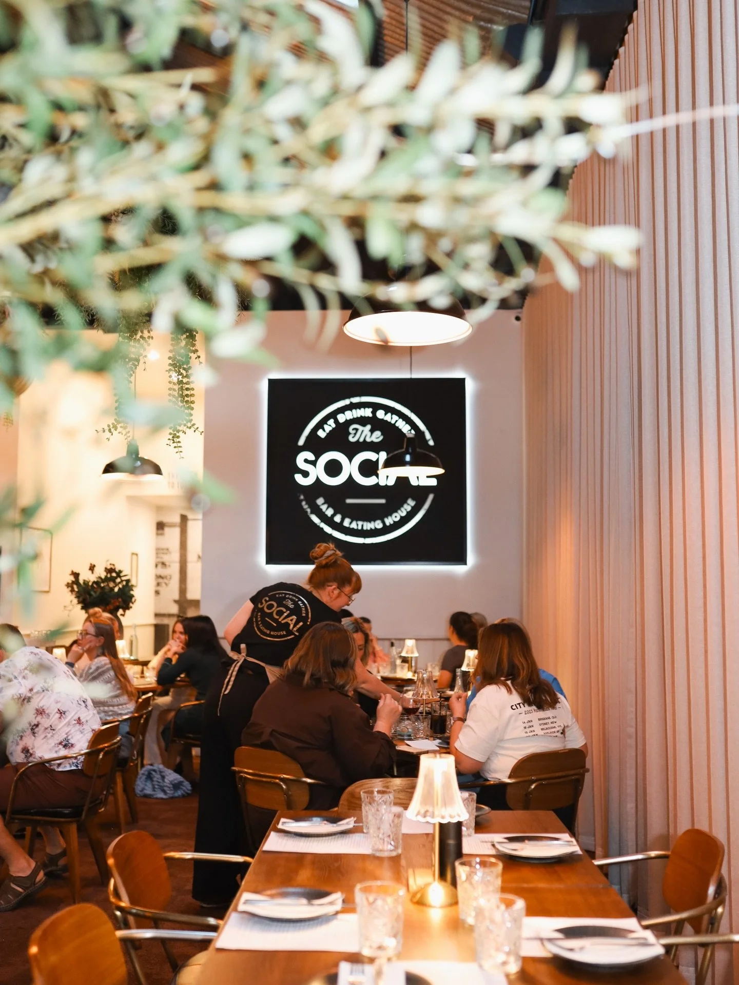 A busy restaurant interior with diners sitting at tables, a server attending to them, and warm lighting, with a lit sign on the wall reading 'The Social'.