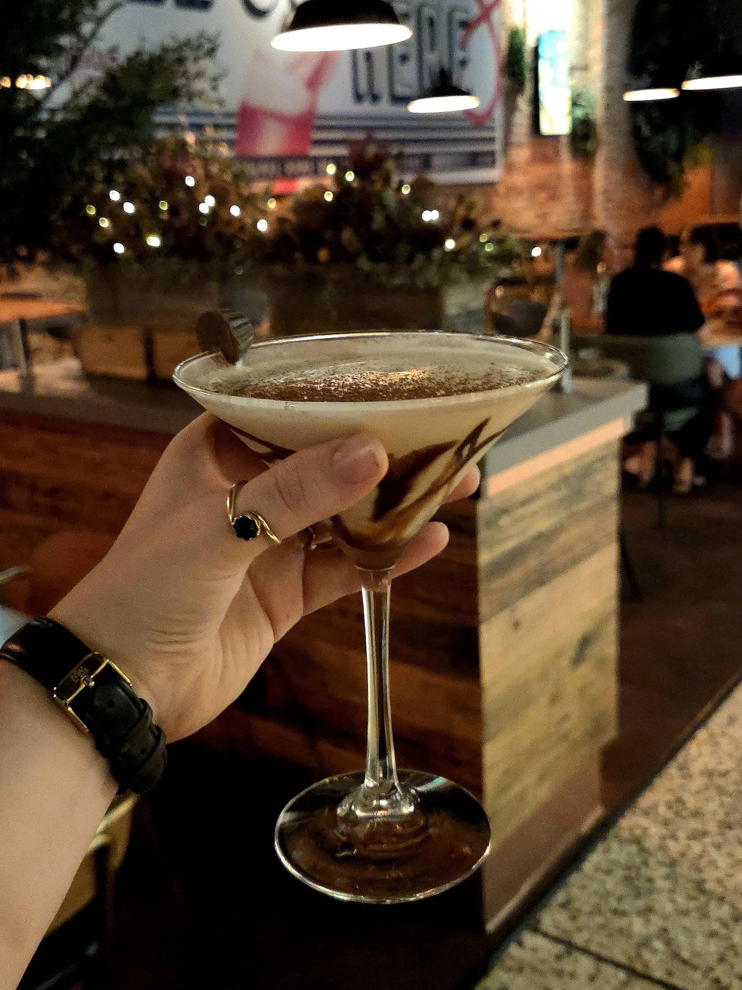 A hand holding a cocktail glass with a dark drink, garnished with a chocolate piece, in a dimly lit restaurant or bar setting.