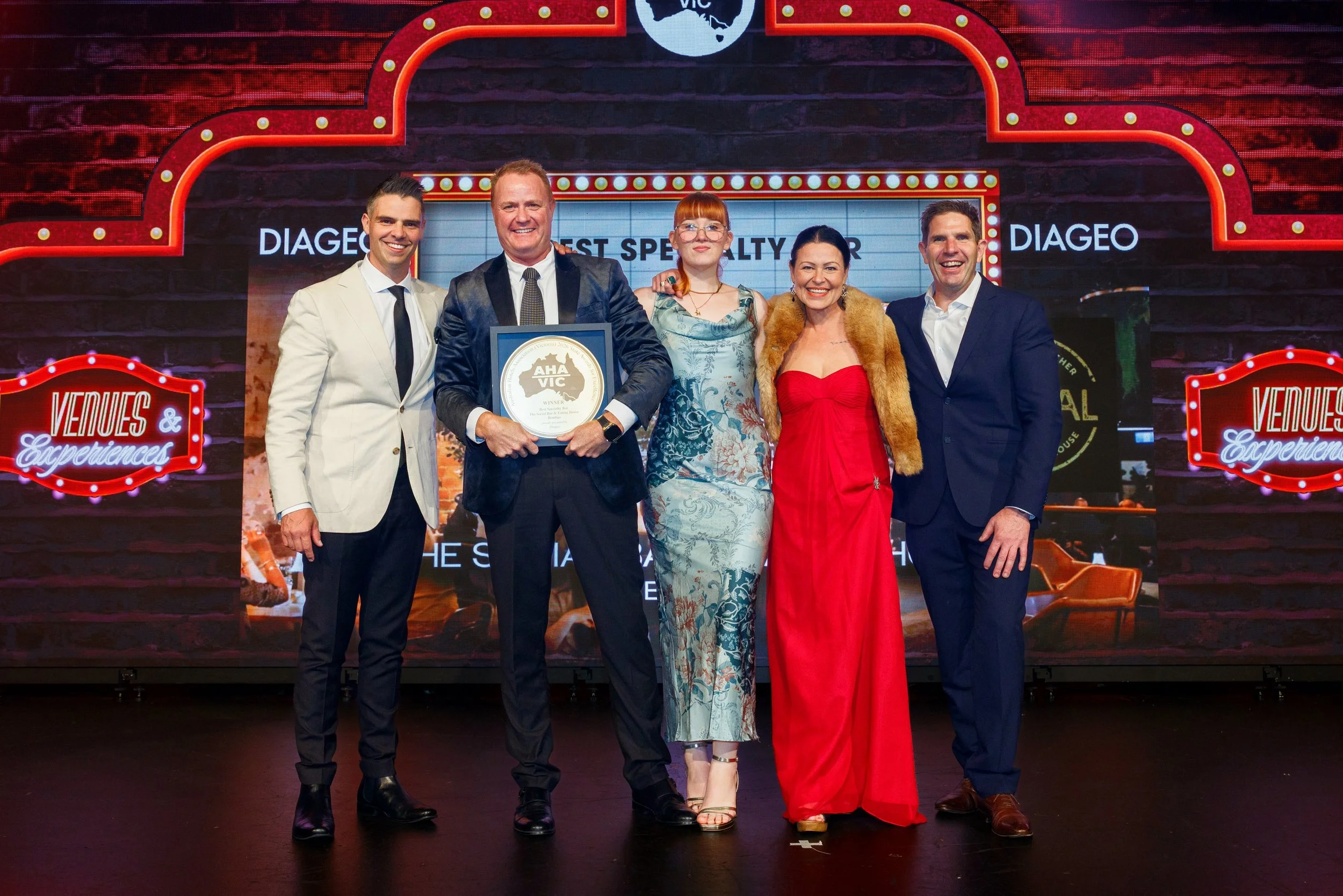 A group of five people standing together on a stage with a neon sign background, celebrating an award win at an event. One man holds a framed certificate. The group is dressed in formal attire and smiling.