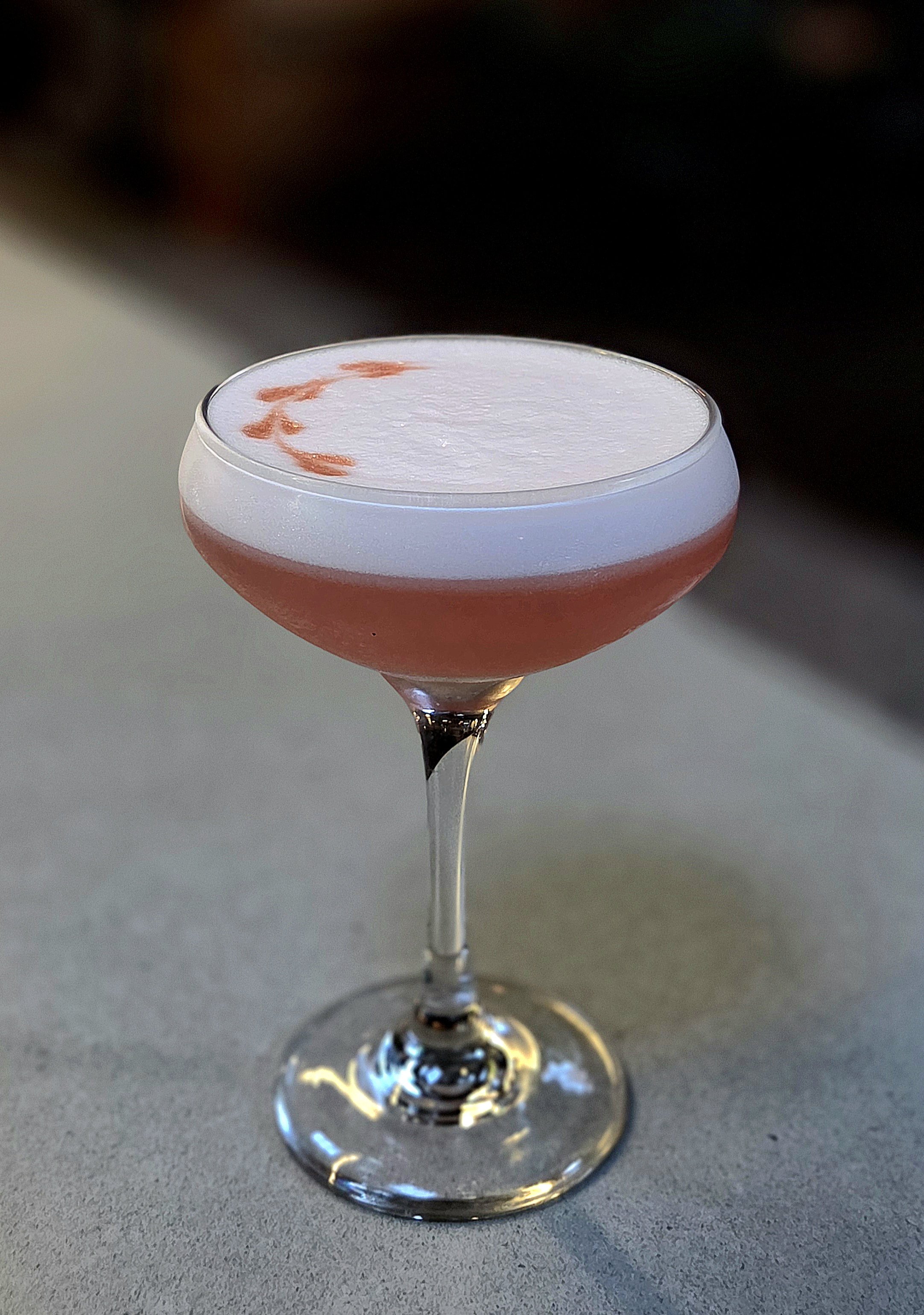 A cocktail glass with a red and white layered drink, adorned with a white foam top and a small red design on top, placed on a gray surface.
