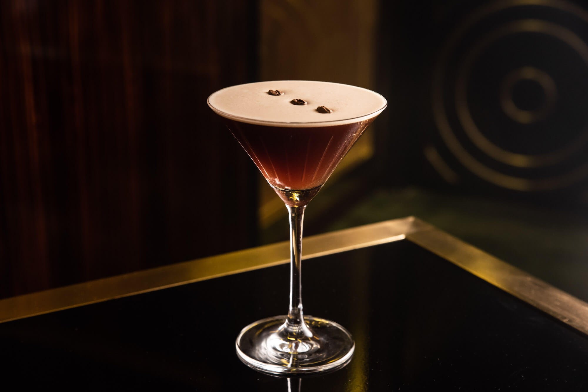 A martini glass filled with a dark cocktail, topped with a thick layer of foam and garnished with three coffee beans, against a dark background.