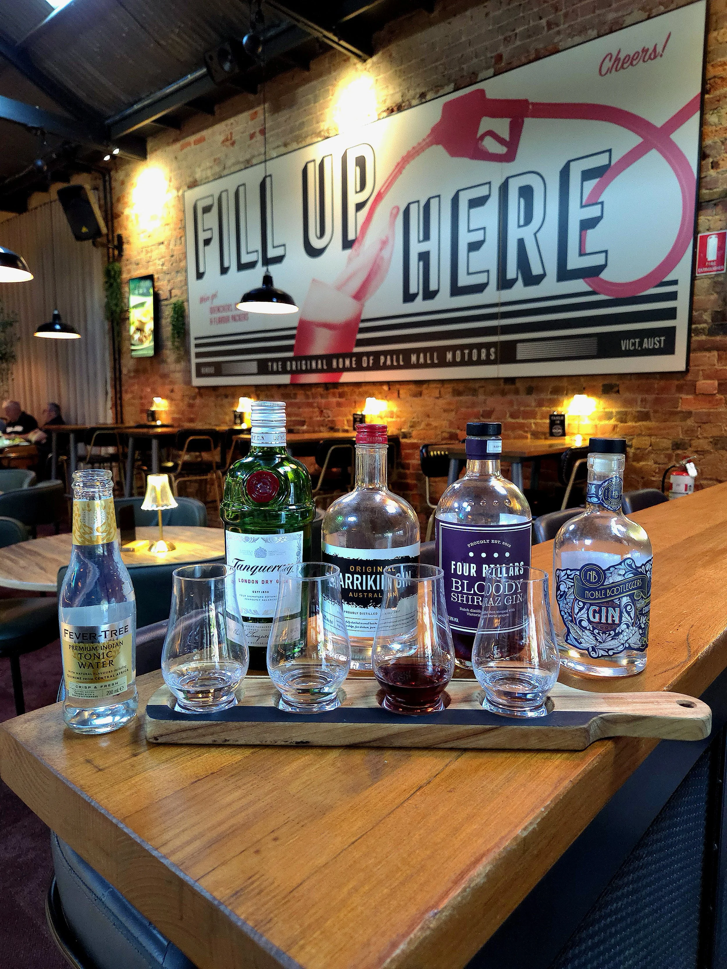A bar tabletop with various bottles of alcohol, including tonic water, gin, and bloody shiraz, and several empty glasses. In the background, a large mural on a brick wall reads 'Fill Up Here' with an illustration of a pink drink and a straw, and a si