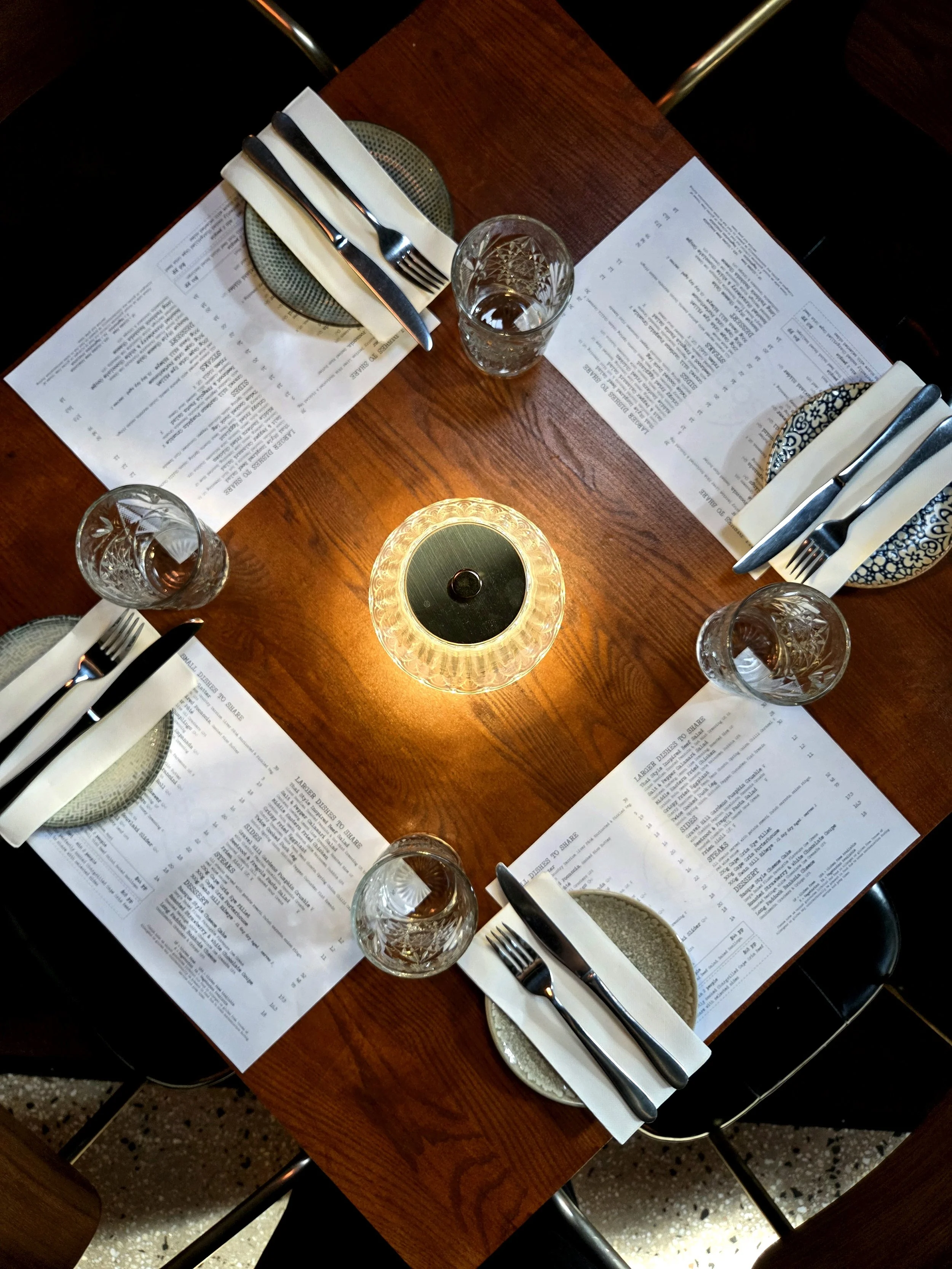A restaurant table setting with four place settings, each with a fork, knife, napkin, and a small plate. There are four glasses of water, paper menus, and a small glowing table lamp in the center.
