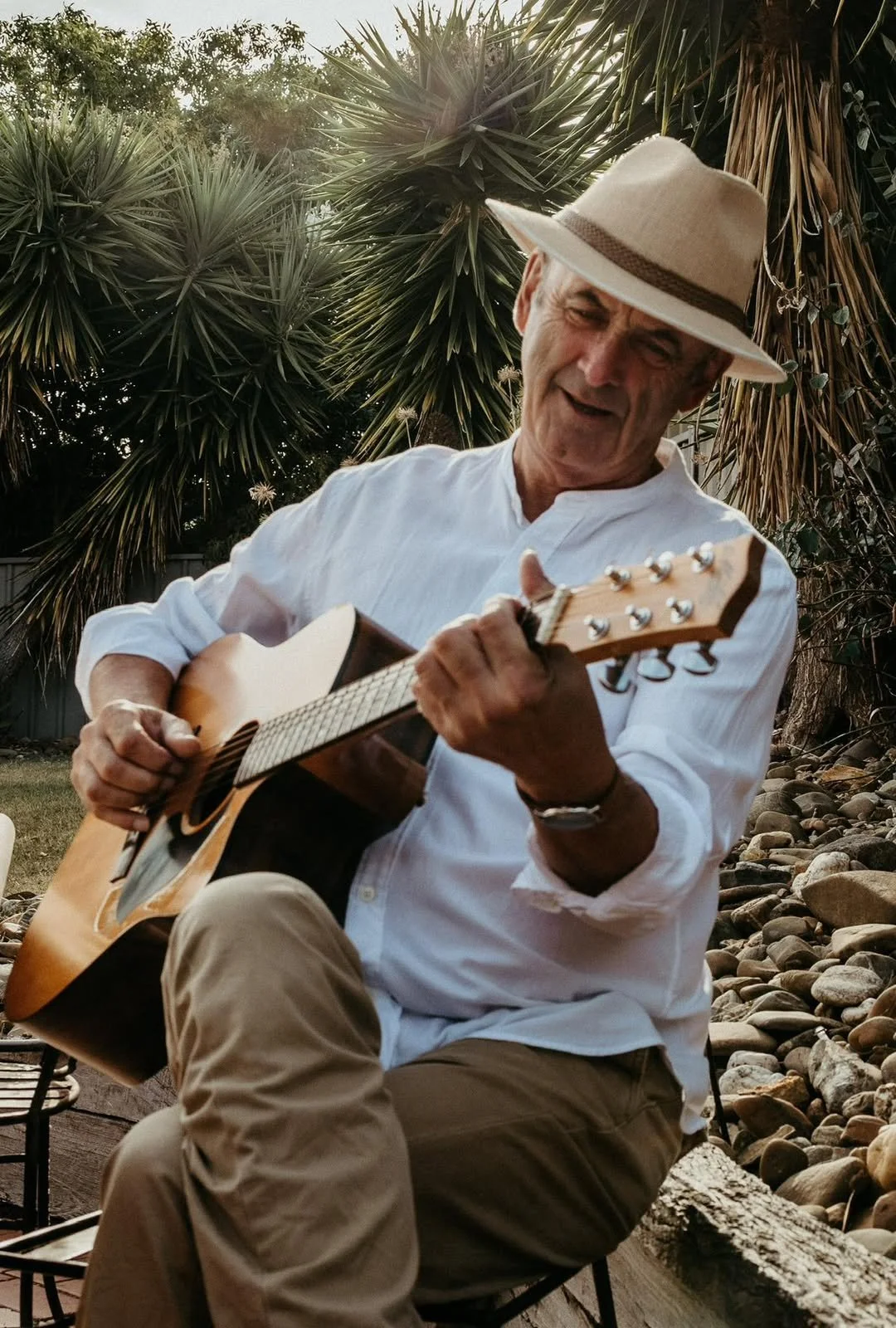 An older man wearing a white shirt and beige pants sits outdoors on a wooden bench, playing an acoustic guitar. He is wearing a wide-brimmed hat and looks happy as he plays. Behind him are large green tropical plants and a rocky ground.