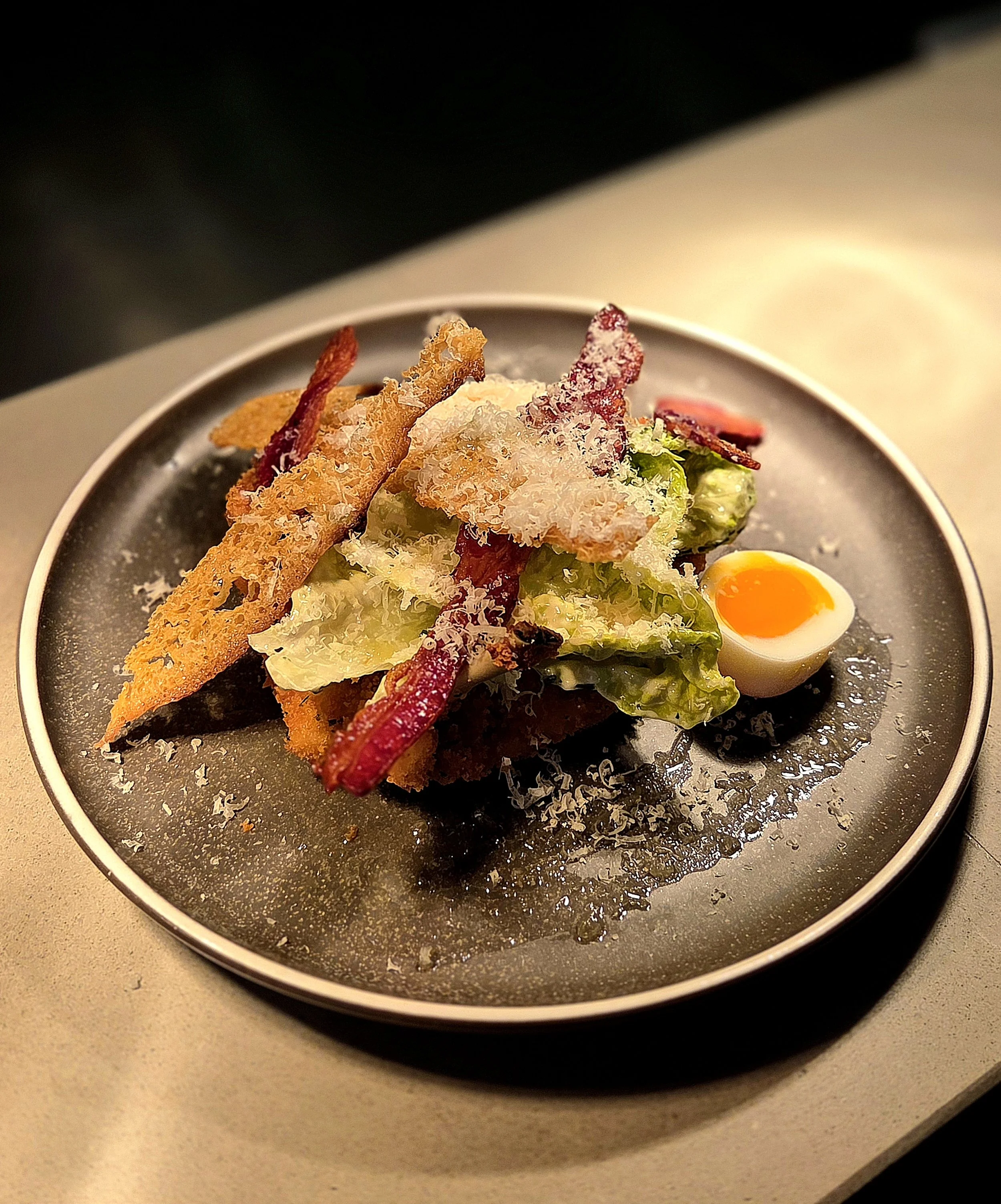 Close-up of a plate with fried chicken strips, shredded cheese, lettuce, sliced tomato, and a halved soft-boiled egg, on a beige countertop.