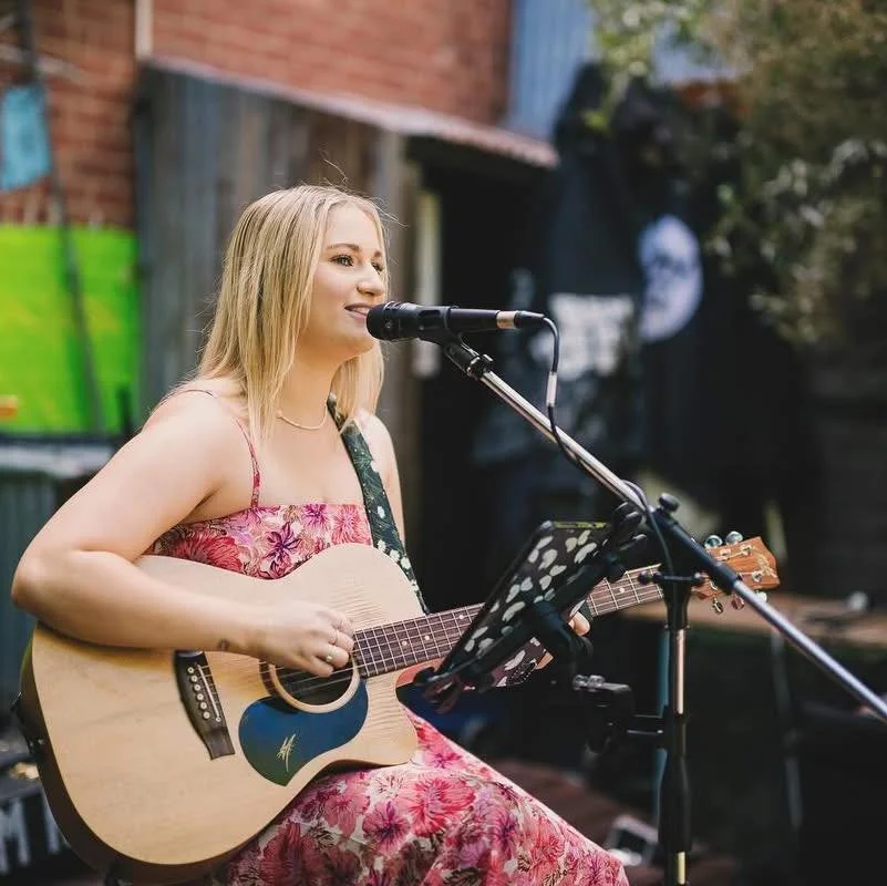Young woman playing acoustic guitar and singing into a microphone outdoors.