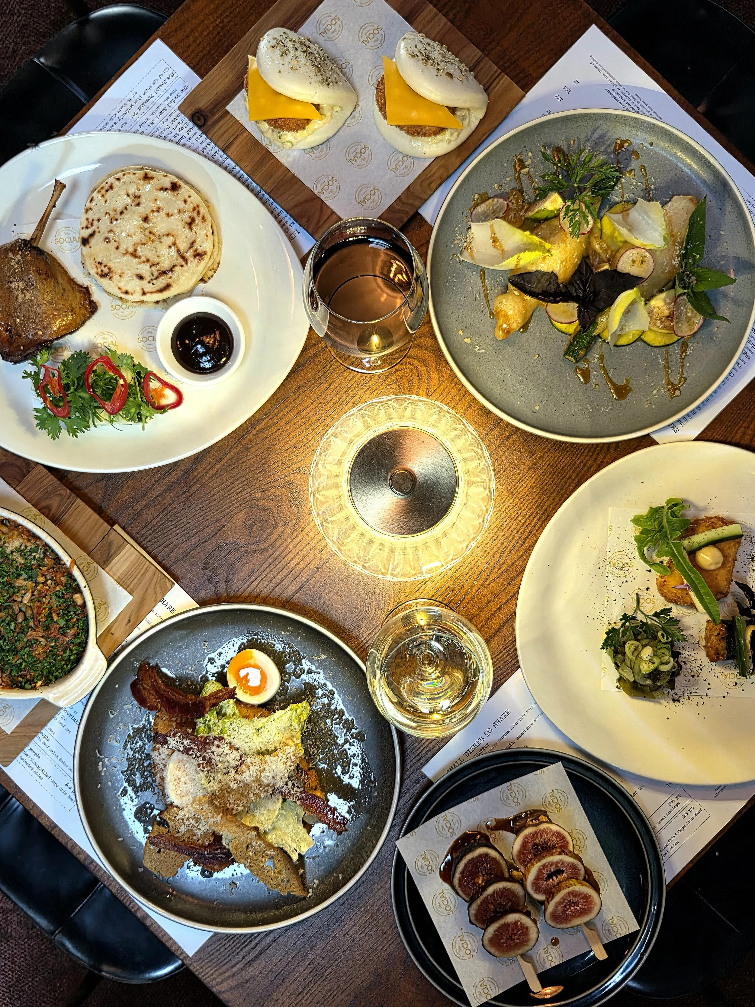 A table with various dishes including sliders, grilled meat, flatbread, salad, and wine, set on a wooden table with sheets of paper underneath.