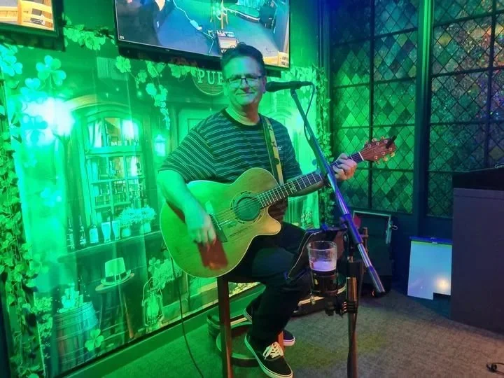 A man with glasses, wearing a striped shirt and jeans, playing an acoustic guitar seated at a microphone in a dimly lit bar or pub with green lighting and shamrock decorations.