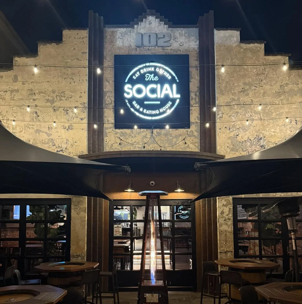 Exterior view of a restaurant called 'The Social' at night, with a large illuminated sign on a rustic brick wall, string lights overhead, and patio seating with tables and chairs.