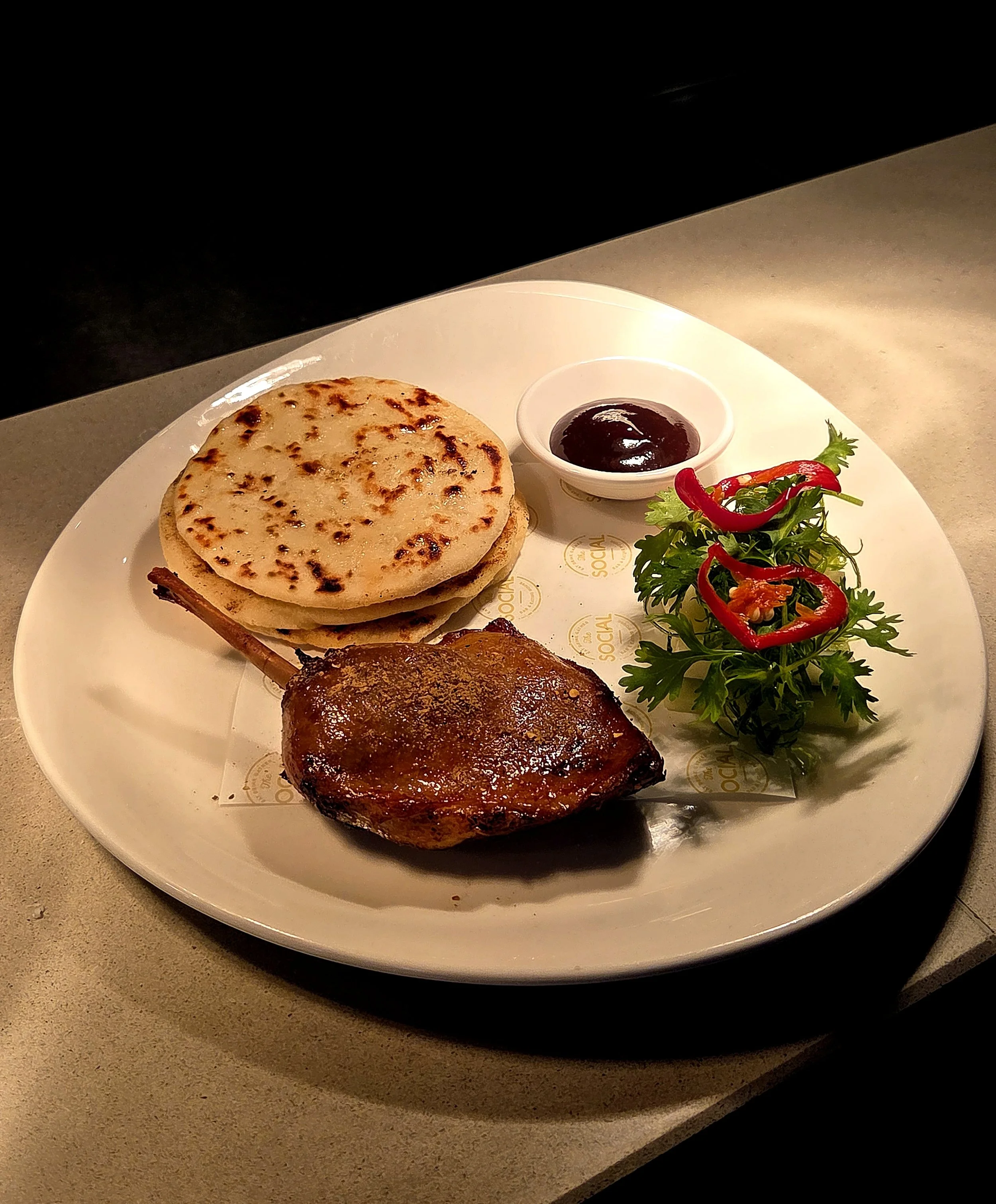 Plate with naan bread, small bowl of dark sauce, grilled piece of meat, and a garnish of fresh parsley and red peppers.