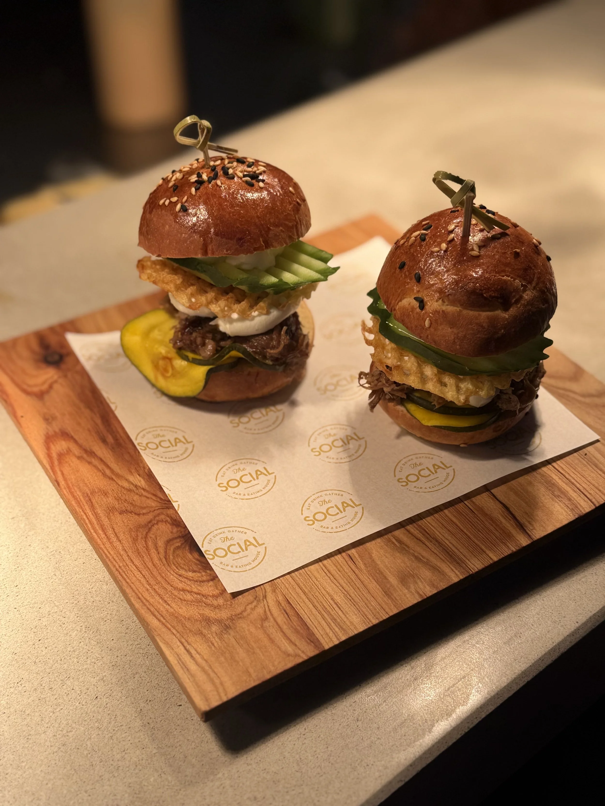 Two mini burgers with various toppings on a wooden serving tray lined with paper that says "The Social".