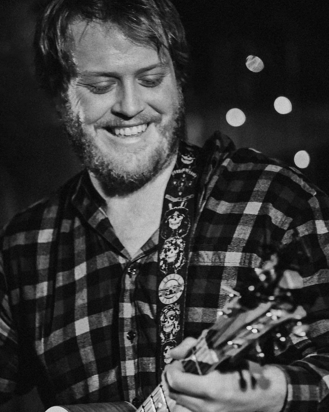 A black and white photo of a smiling man with a beard and mustache, wearing a plaid shirt and holding a guitar, with blurred circular lights in the background.