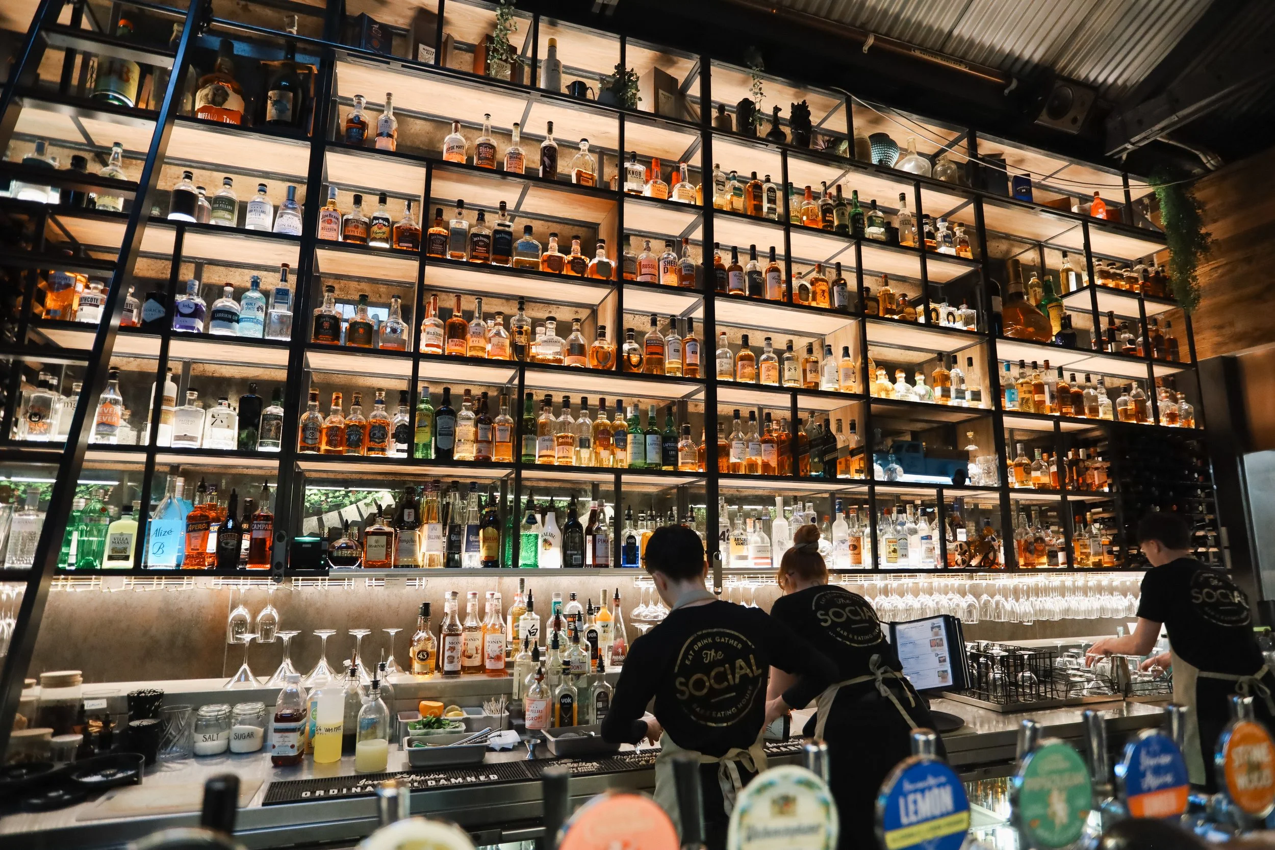 A bar with multiple shelves filled with various liquor bottles behind the counter. There are bar staff working, and the setting appears to be in a restaurant or a bar with a modern industrial design.