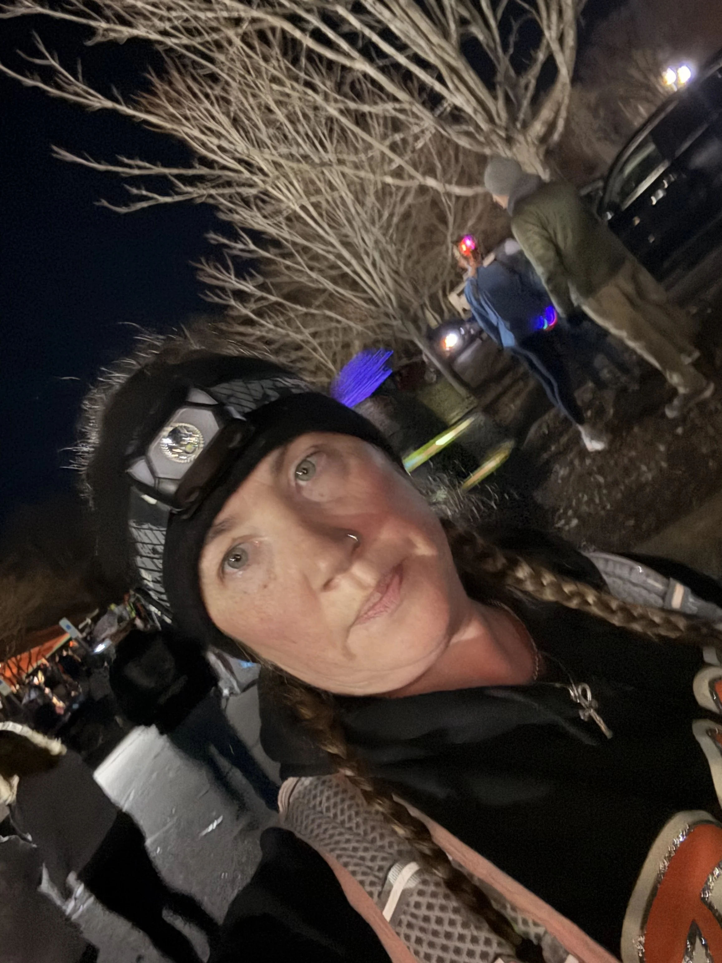 A woman taking a selfie outdoors at night with people and parked cars in the background. She is wearing a headlamp, a black jacket, and has braided hair.