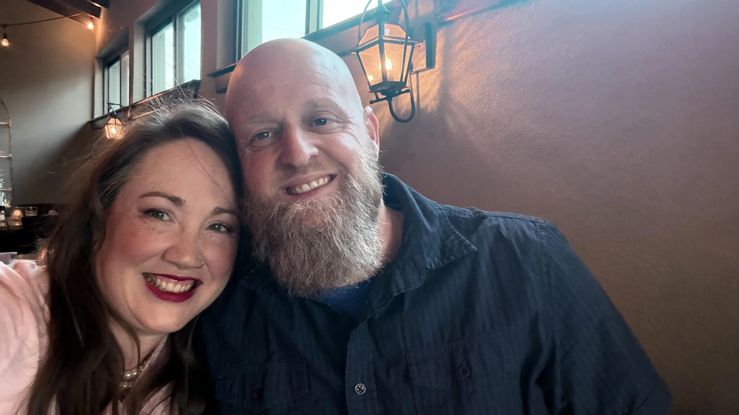 A smiling woman with long brown hair and red lipstick, and a bald man with a beard, smiling together in a restaurant with warm lighting and windows in the background.