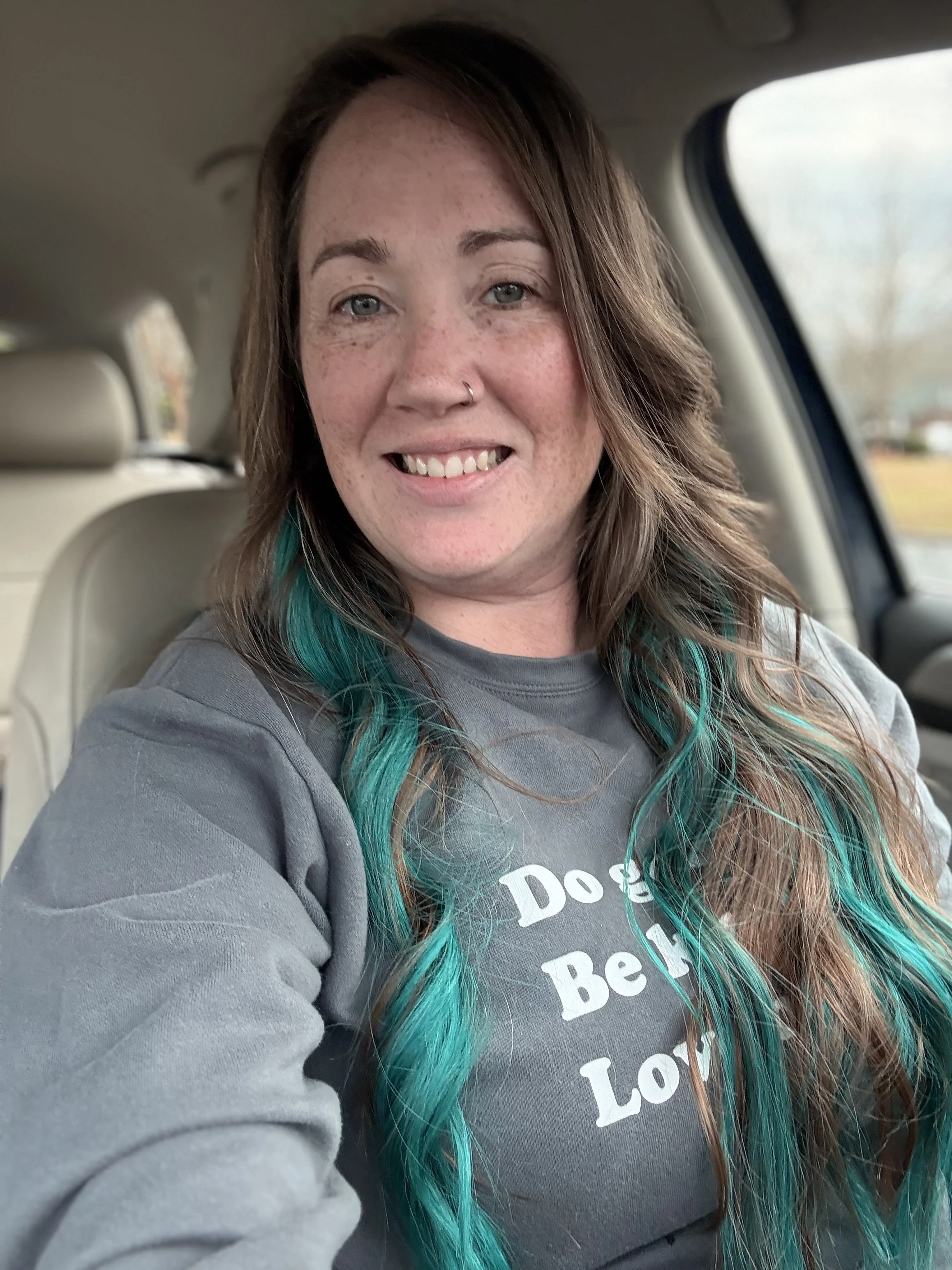 A woman with long wavy brown hair with turquoise streaks, smiling while sitting in a car, wearing a gray shirt with white text.