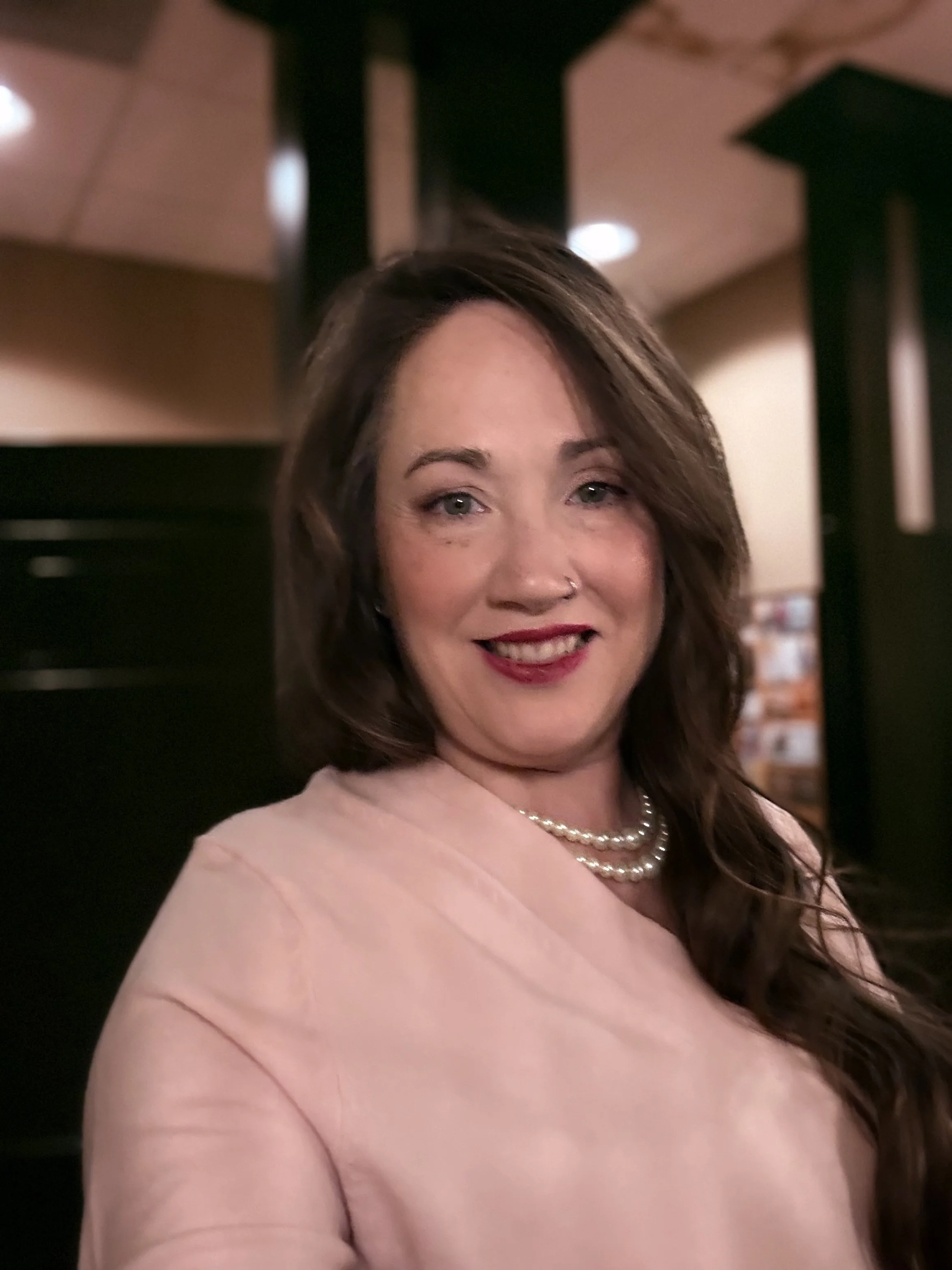 A woman with brown hair, light makeup, red lipstick, a pearl necklace, and a pinkish-beige top, smiling in an indoor setting.