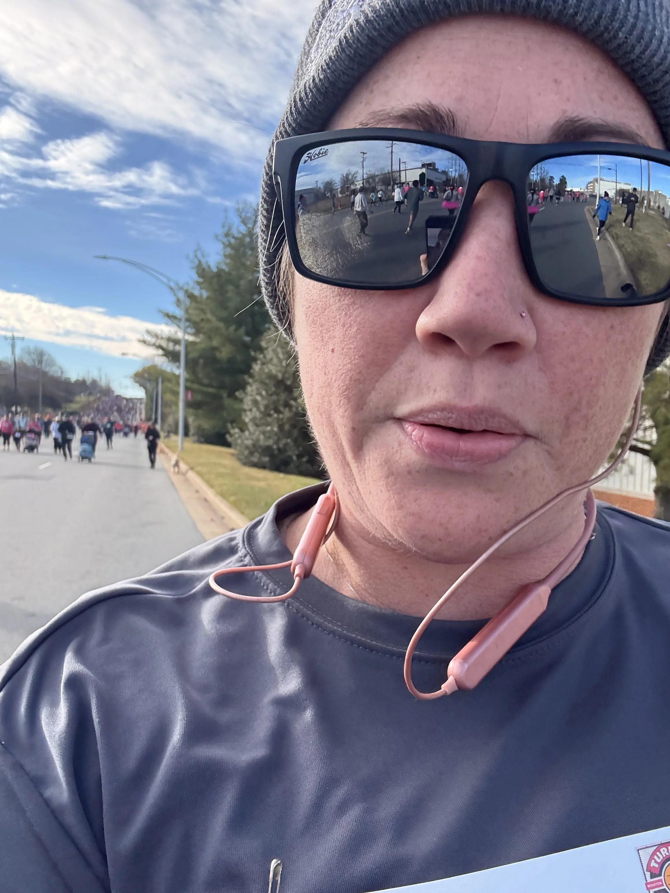 A woman taking a selfie during a running event, with a crowd of runners in the background and her reflection visible in her sunglasses.