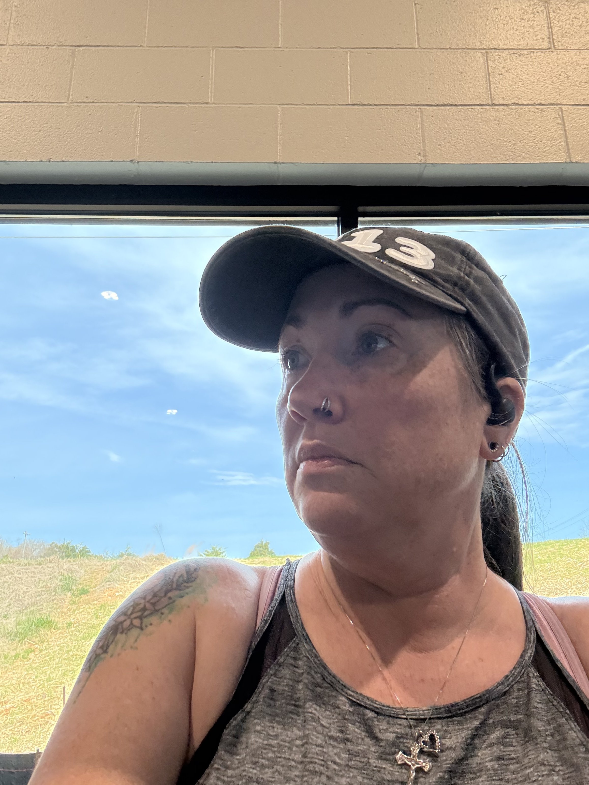 Woman wearing a black cap, grey tank top, and a cross necklace, sitting indoors near a large window with a view of blue sky and green landscape outside.