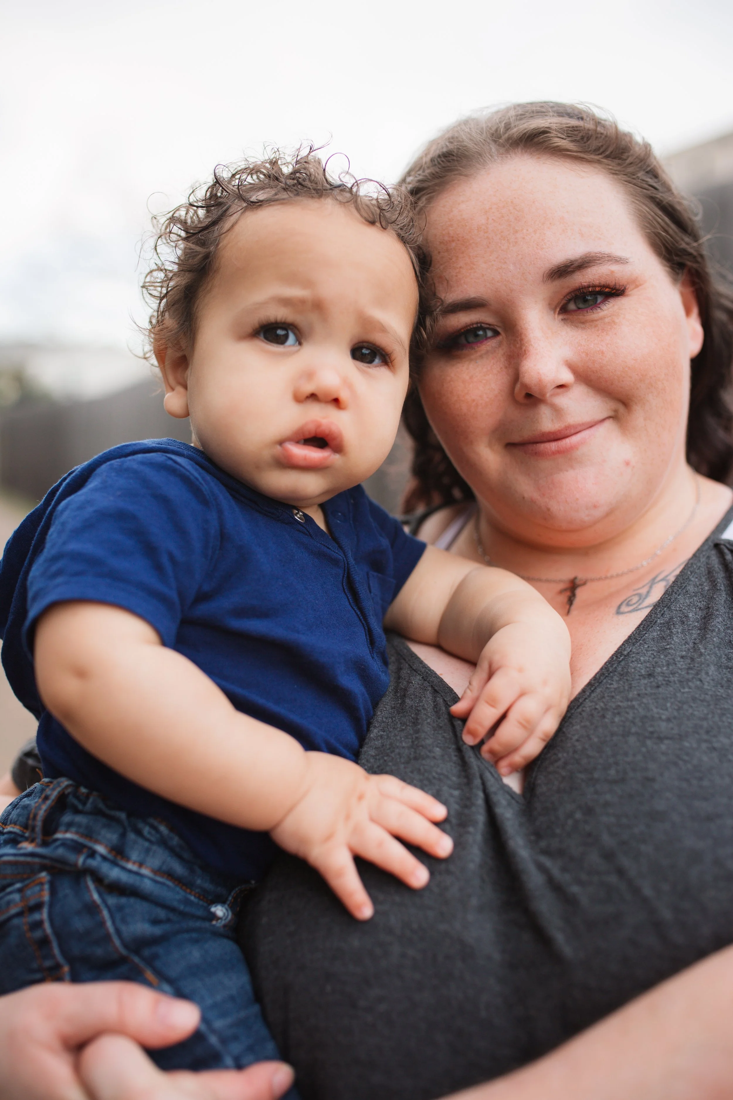 A woman with light skin and freckles holding a young boy with curly hair and tan skin, both smiling gently outdoors on a cloudy day.
