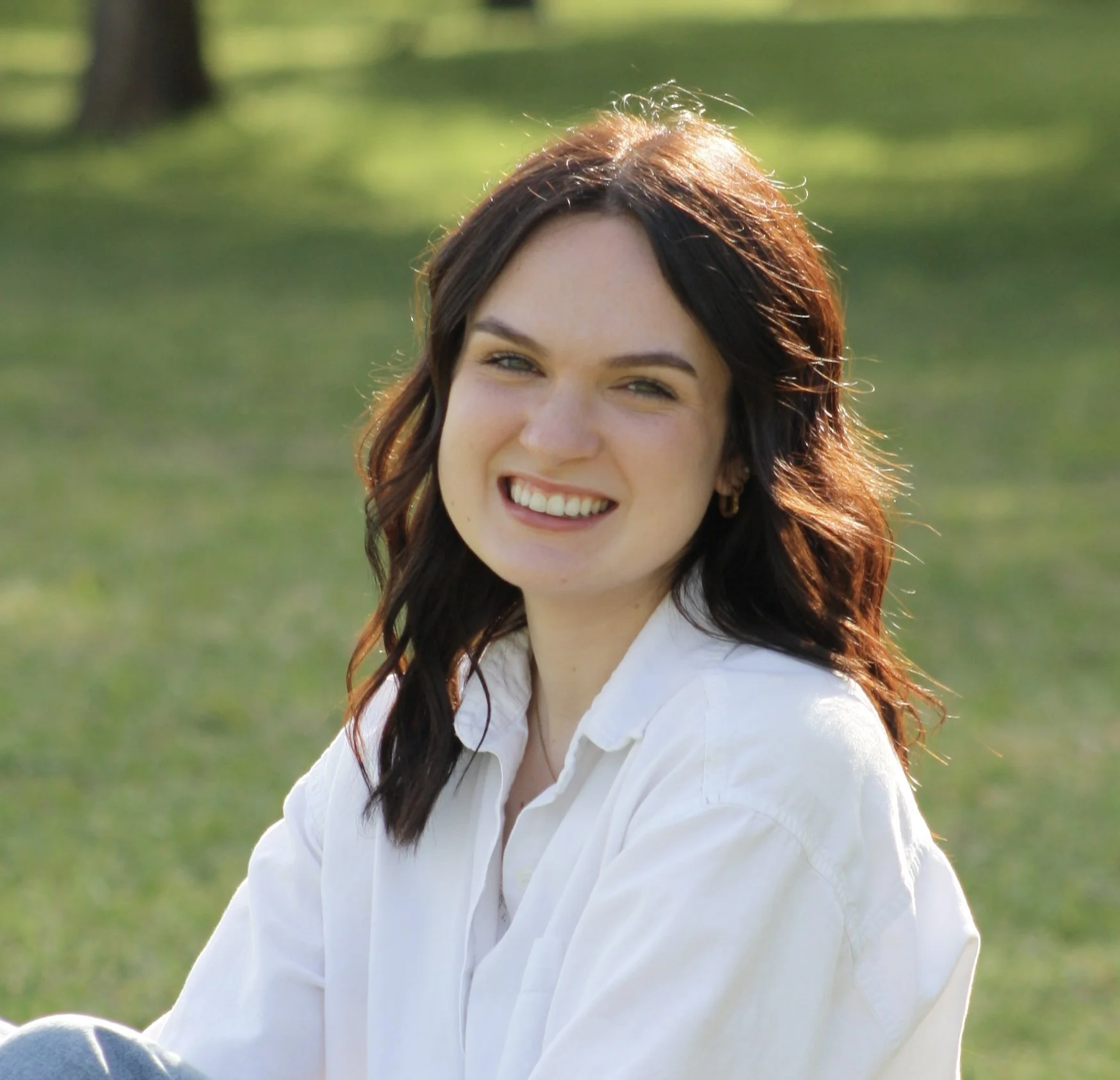 A young woman with long, wavy brown hair, smiling outdoors on a sunny day, wearing a white button-up shirt, sitting on the grass in a park.