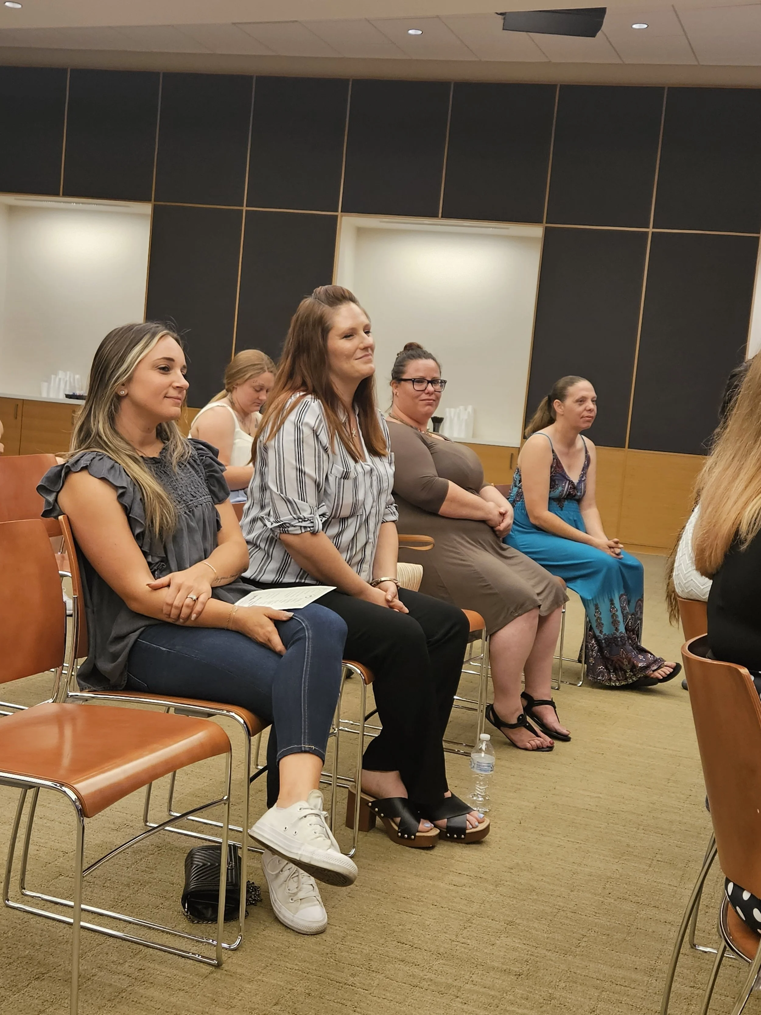 Women sitting in chairs in a room with wood-paneled walls, engaged in an event or presentation.