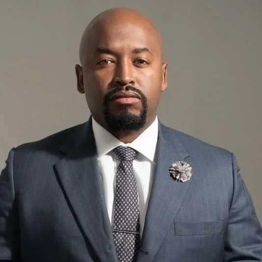 A bald Black man wearing a navy blue suit, white shirt, and patterned tie, with a flower-shaped lapel pin.