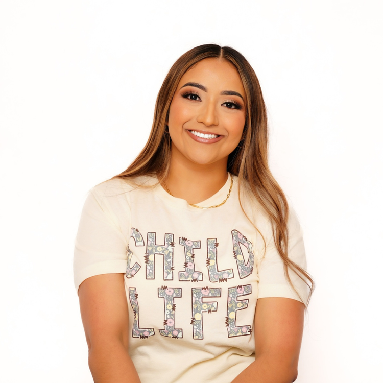 A smiling woman with long brown hair, wearing a white T-shirt with colorful floral letters spelling 'CHILD LIFE,' stands against a plain white background.