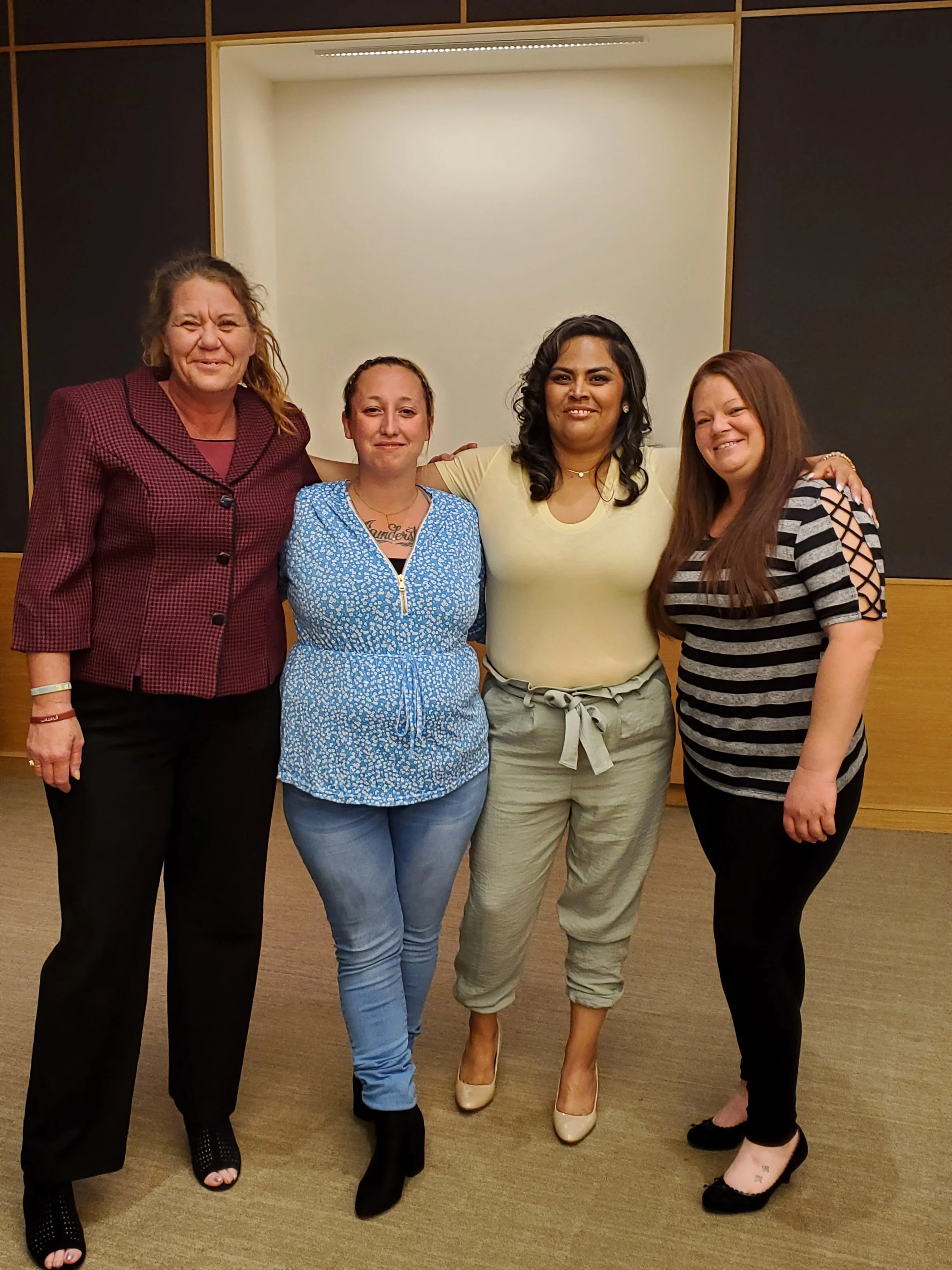 Four women standing together indoors for a photo, smiling and posing. The background features a cream-colored wall and black panels.