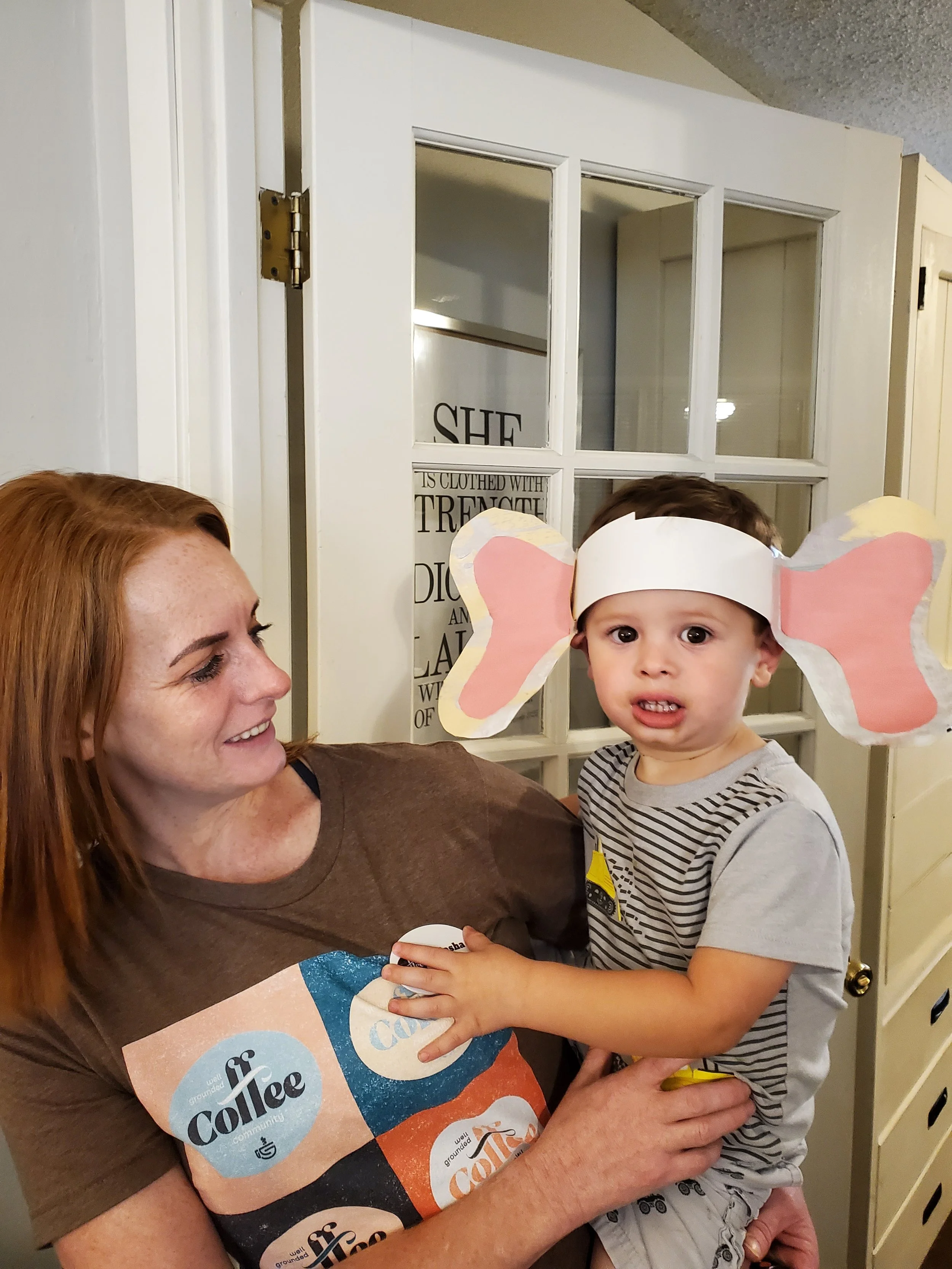 A woman with red hair and a young boy with dinosaur paper wings on his head are smiling and looking at each other inside a home. The woman is holding a sign with the words "grounded coffee community" on it. There is a white door with glass panes in the background.