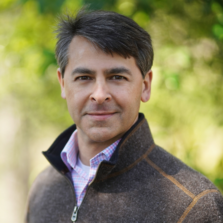 A man with short dark hair and light skin, wearing a dark brown zip-up jacket over a pink and blue checkered shirt, outdoors with a blurred green background.