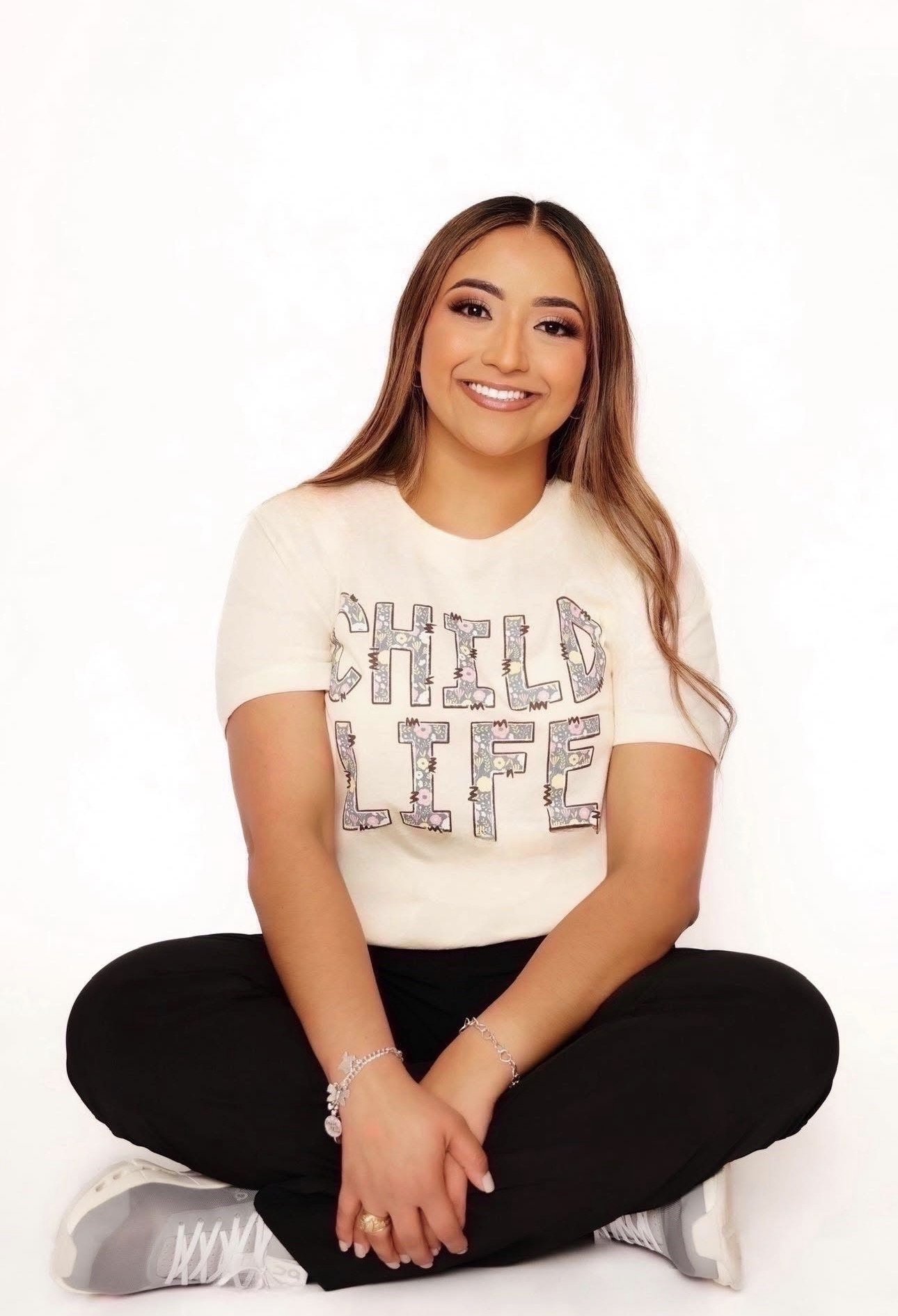 A smiling woman with long brown hair, wearing a white T-shirt with colorful floral letters spelling 'CHILD LIFE,' stands against a plain white background.