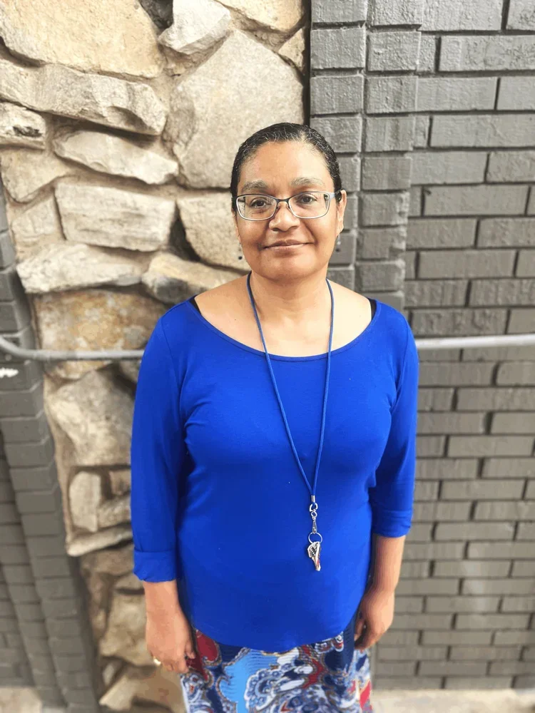 A woman with glasses and short dark hair stands in front of a stone and brick wall, wearing a blue top and a patterned skirt, with a lanyard around her neck.