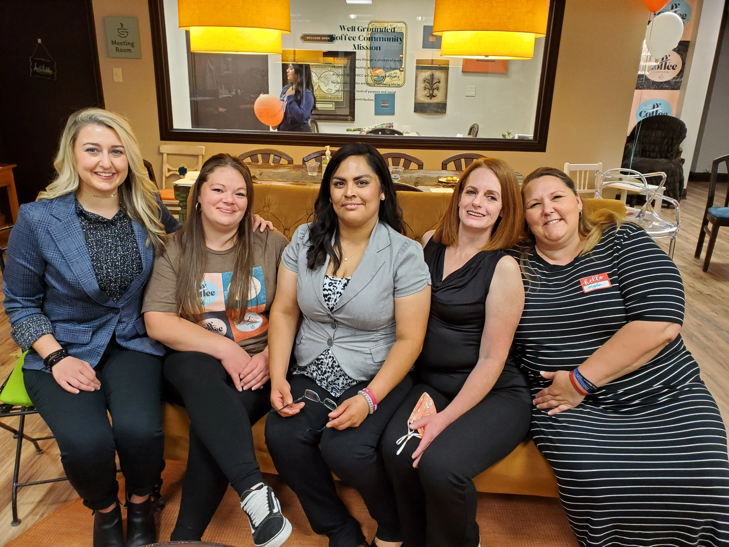 Five women sitting closely together in a cozy coffee shop, smiling at the camera. Behind them, there is a large window with a reflection of a woman holding a balloon, and signs on the wall, including one that says 'Well Grounded Coffee Community Mission.' The women are dressed casually, and some are wearing name tags.