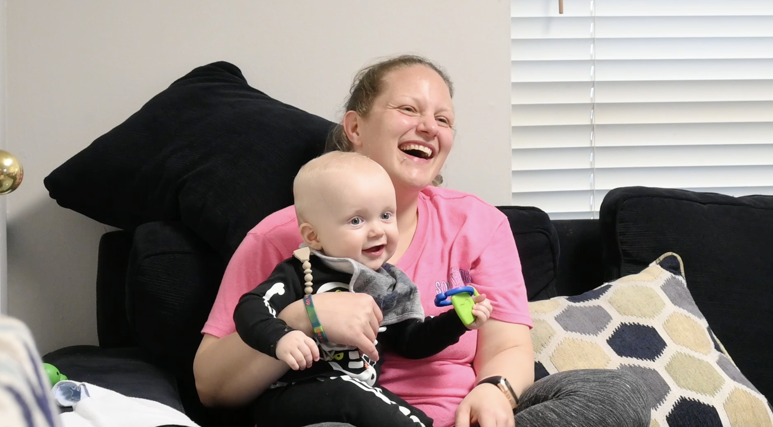 A woman and a young child sitting on a black couch in a living room, smiling and enjoying each other's company. The woman is wearing a pink shirt and the child is dressed in a skeleton costume, holding a toy and wearing a colorful wristband.