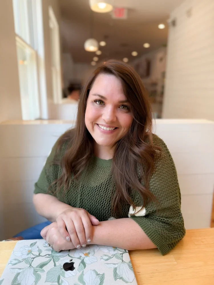 A young woman with long brown hair, smiling, sitting at a table in a cozy cafe. She is wearing a green knitted sweater, with an Apple device and a floral napkin on the table in front of her.