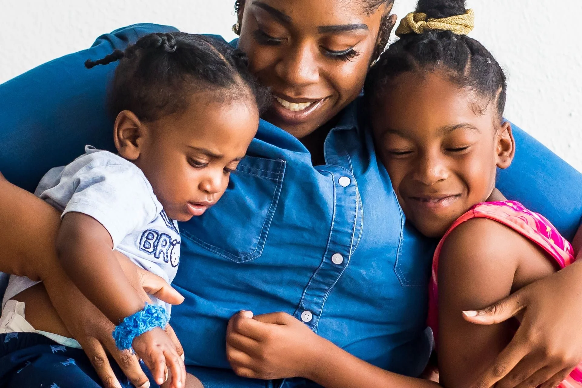 A woman with dark hair and earrings is hugging two young girls, one with braided hair and the other with curly hair and a gold hair tie, both with closed eyes and smiling, against a plain white wall.