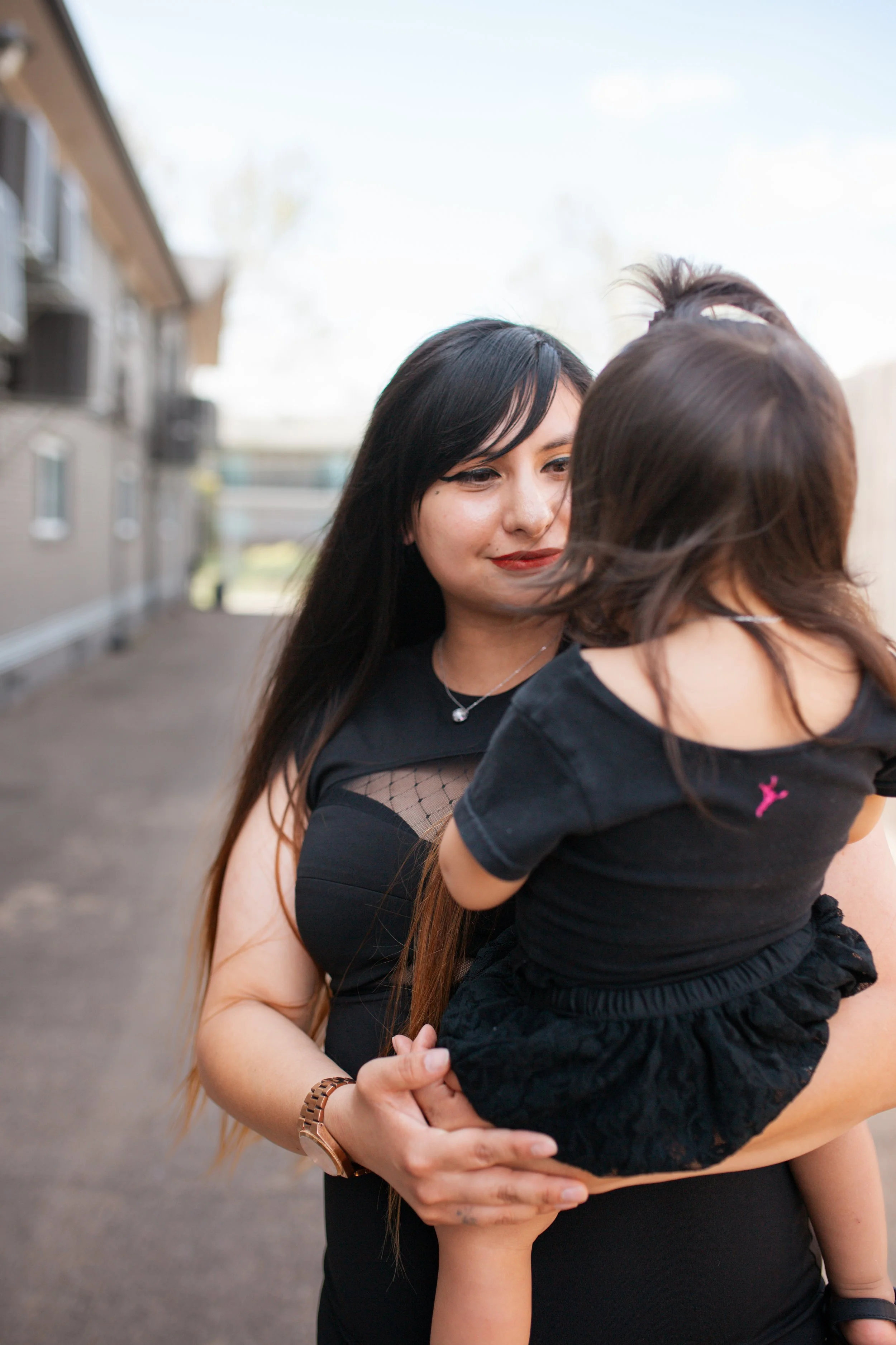 A woman with long dark hair, wearing a black shirt with sheer sleeves, holding a young girl with brown hair, black outfit, and a white bow in her hair, outdoors in front of a residential building.